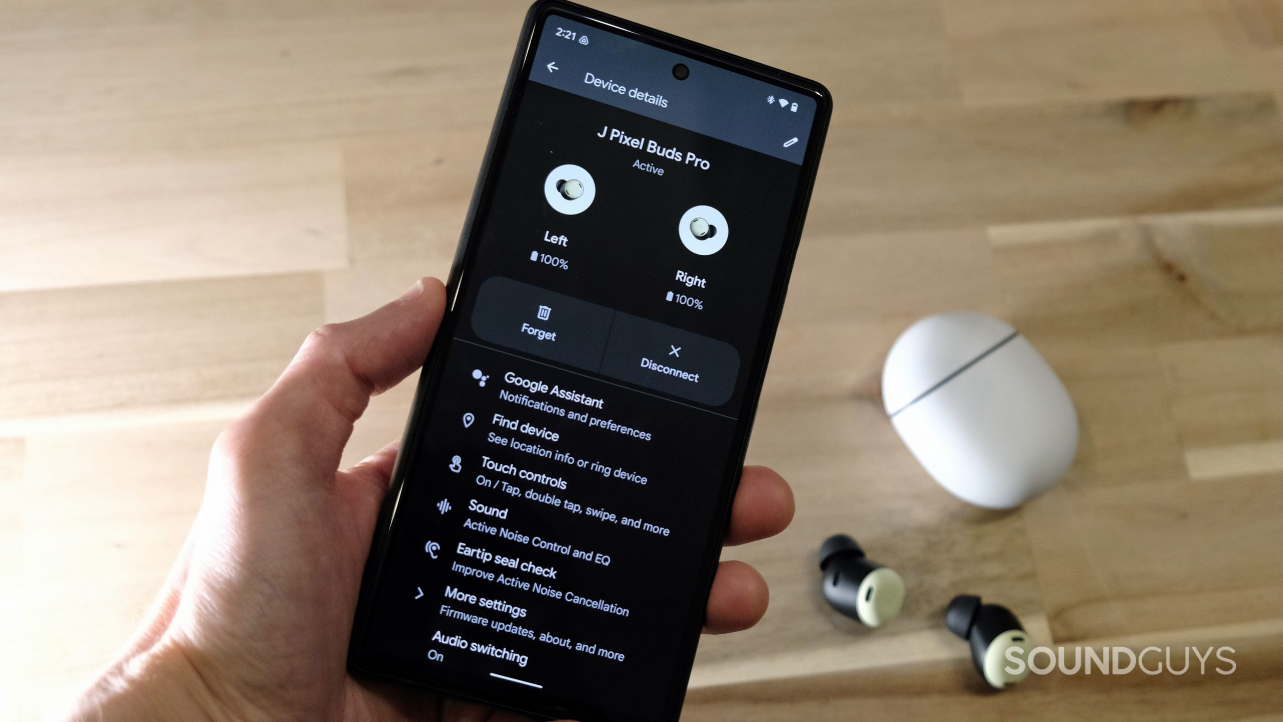 A hand holds a Google Pixel 6 displaying the Google Pixel Buds Pro controls in the Settings app, with the true wireless earbuds on a wooden table in the background.