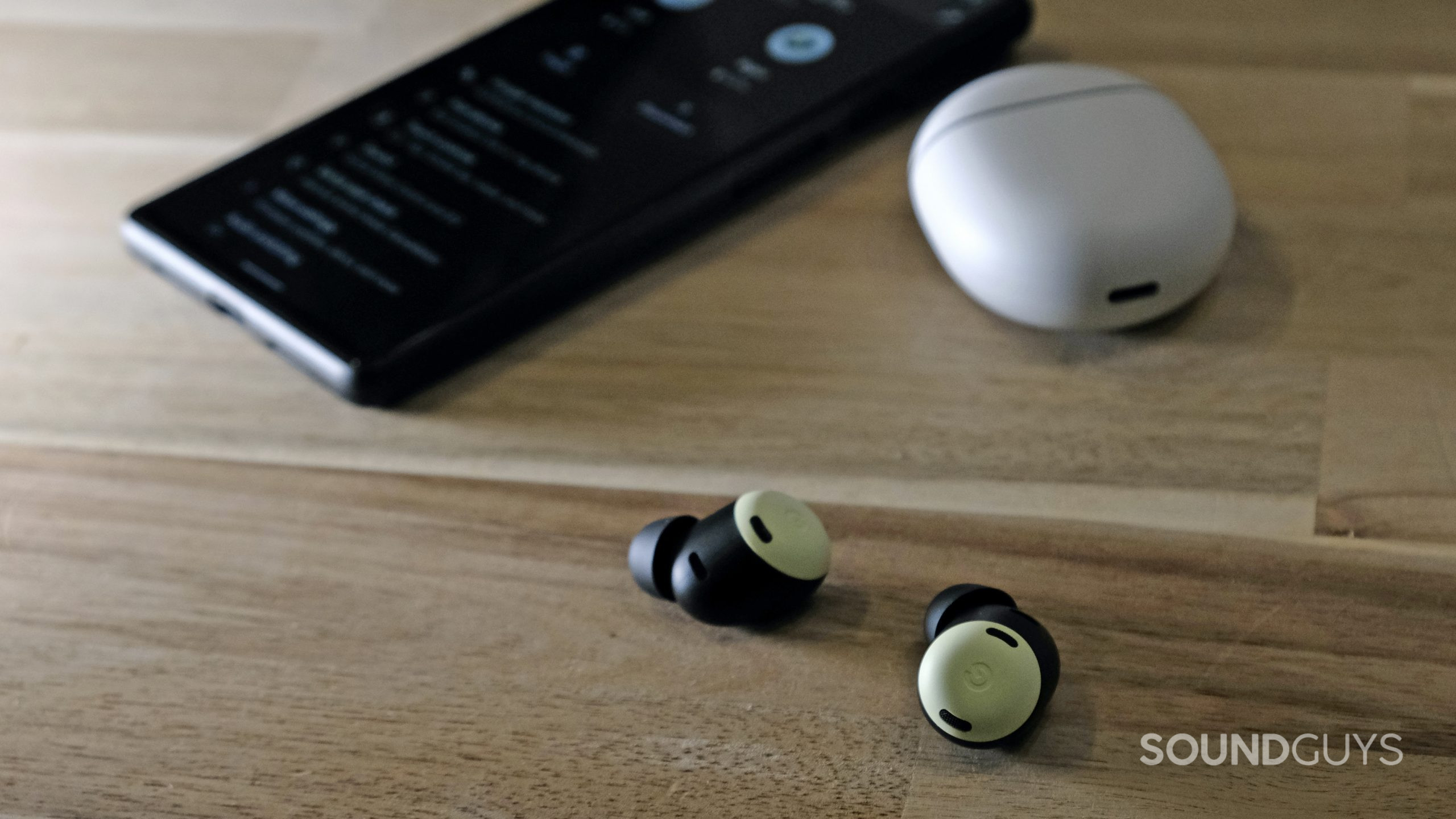 The Google Pixel Buds Pro lays on a wooden table with a Google Pixel 6 behind it.