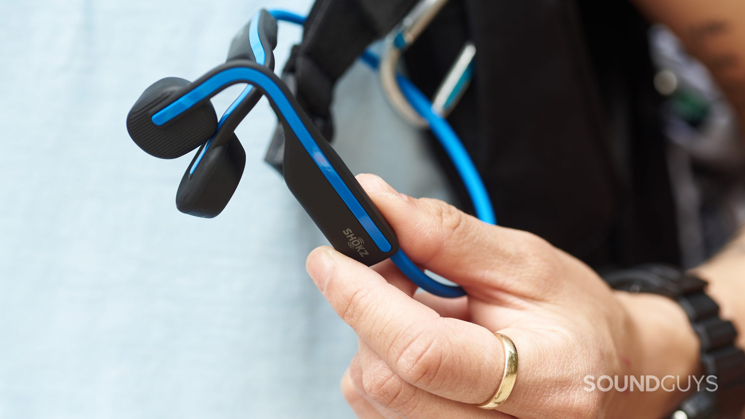 A hand holds the Shokz OpenMove bone conduction headphones in blue before removing the headphones from a carabiner.