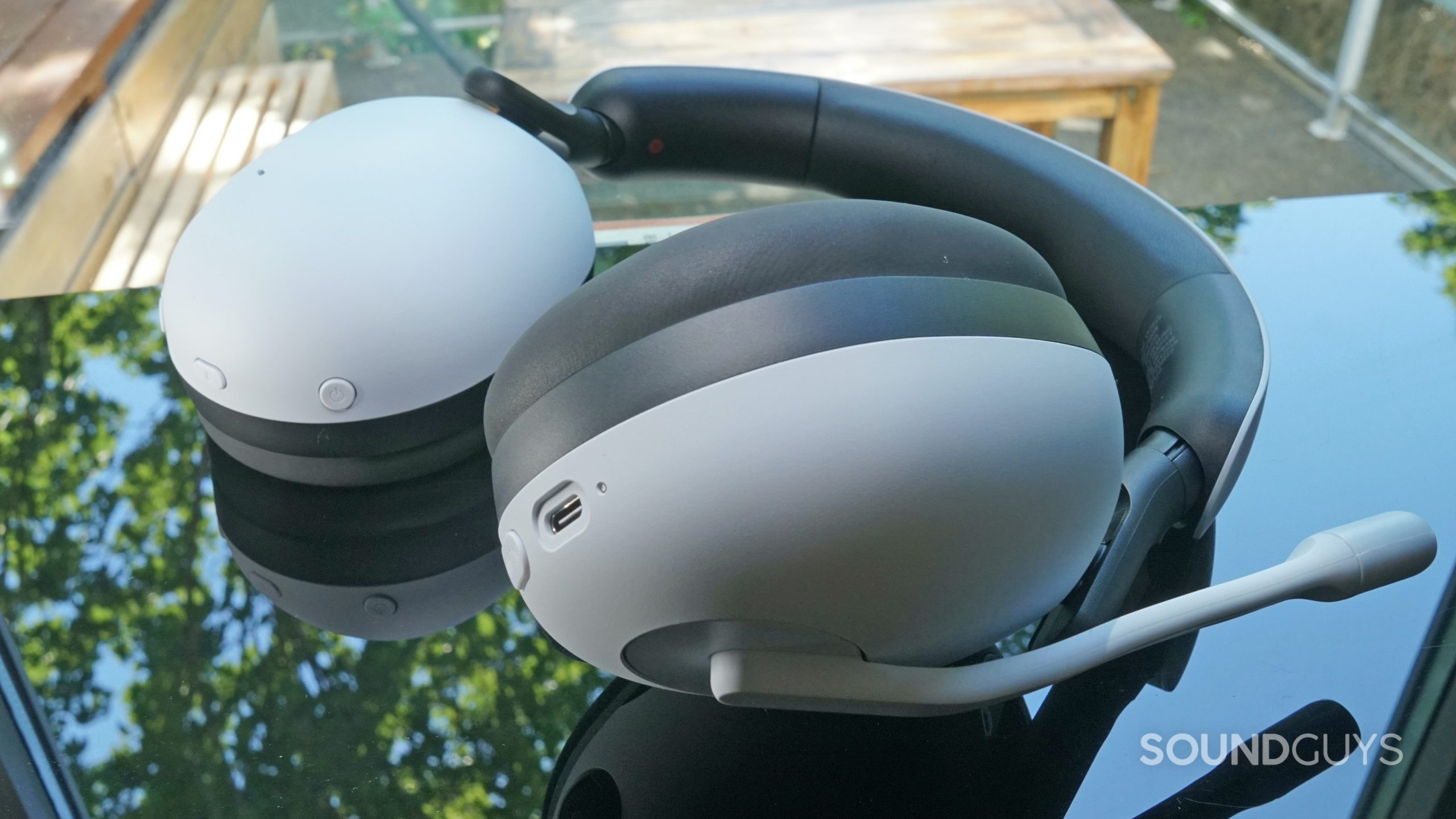 The Sony Inzone H9 gaming headset lays on a reflective surface next to a window on a sunny day. Charging port and buttons are visible