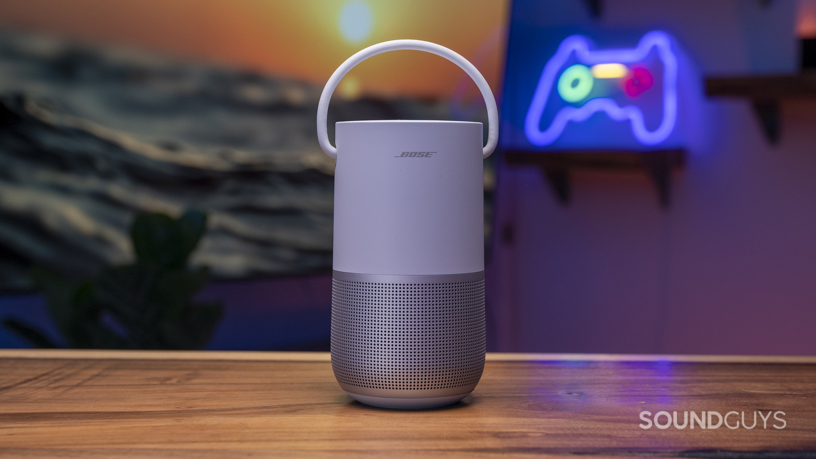 A white Bose Portable Smart Speaker sitting on a table in front of a painting and a neon sign of a controller in the background.