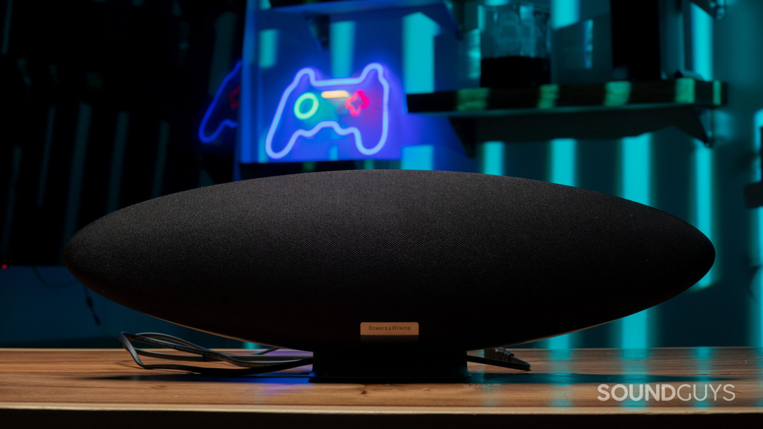 A Bowers &amp; Wilkins Zeppelin resing on a table seen from the front against a background of a neon sign of a game console controller.