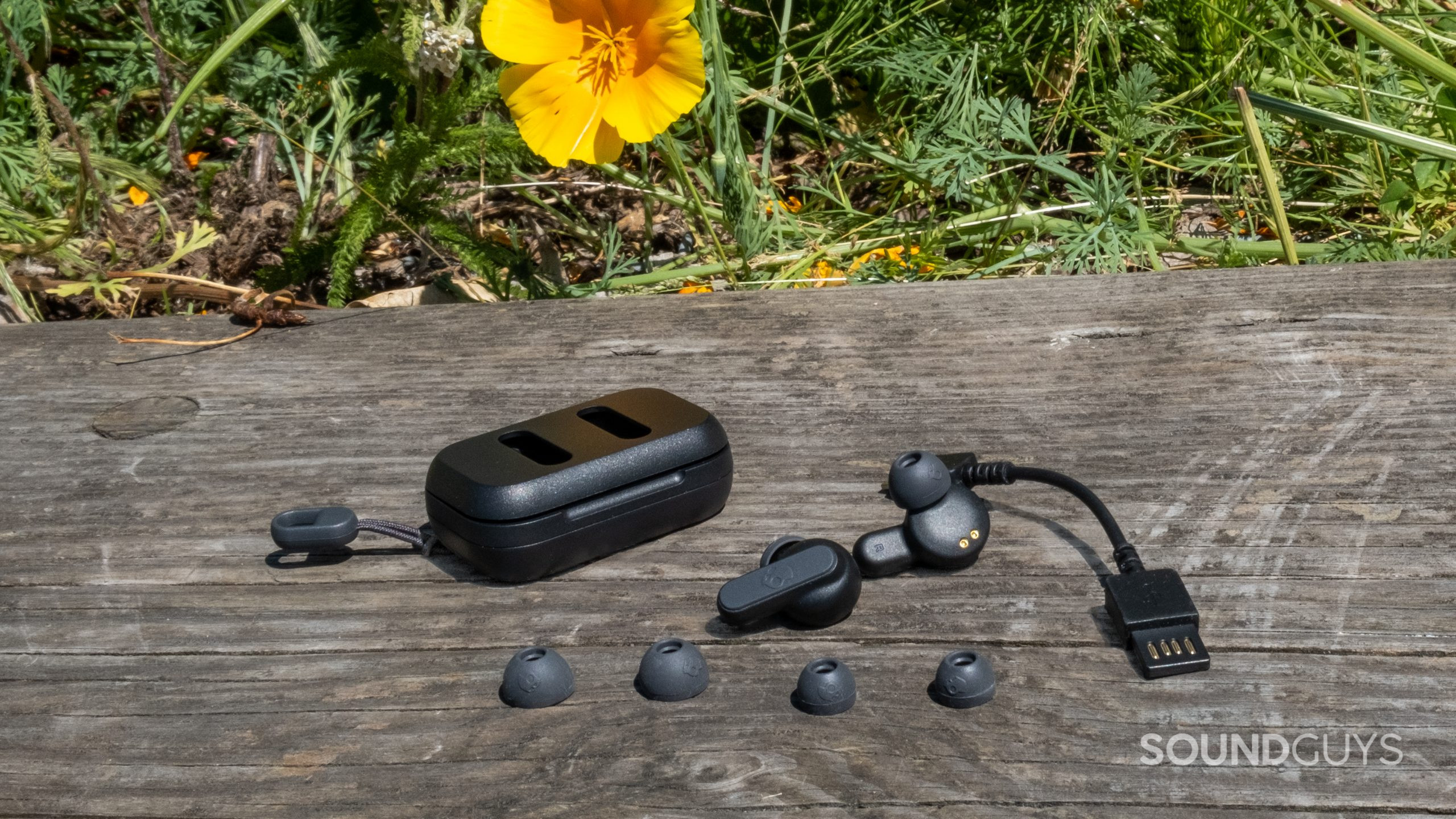 On a wood surface with grass and flowers in the background, the Skullcandy Dime 2 is spread out showing its included accessories.