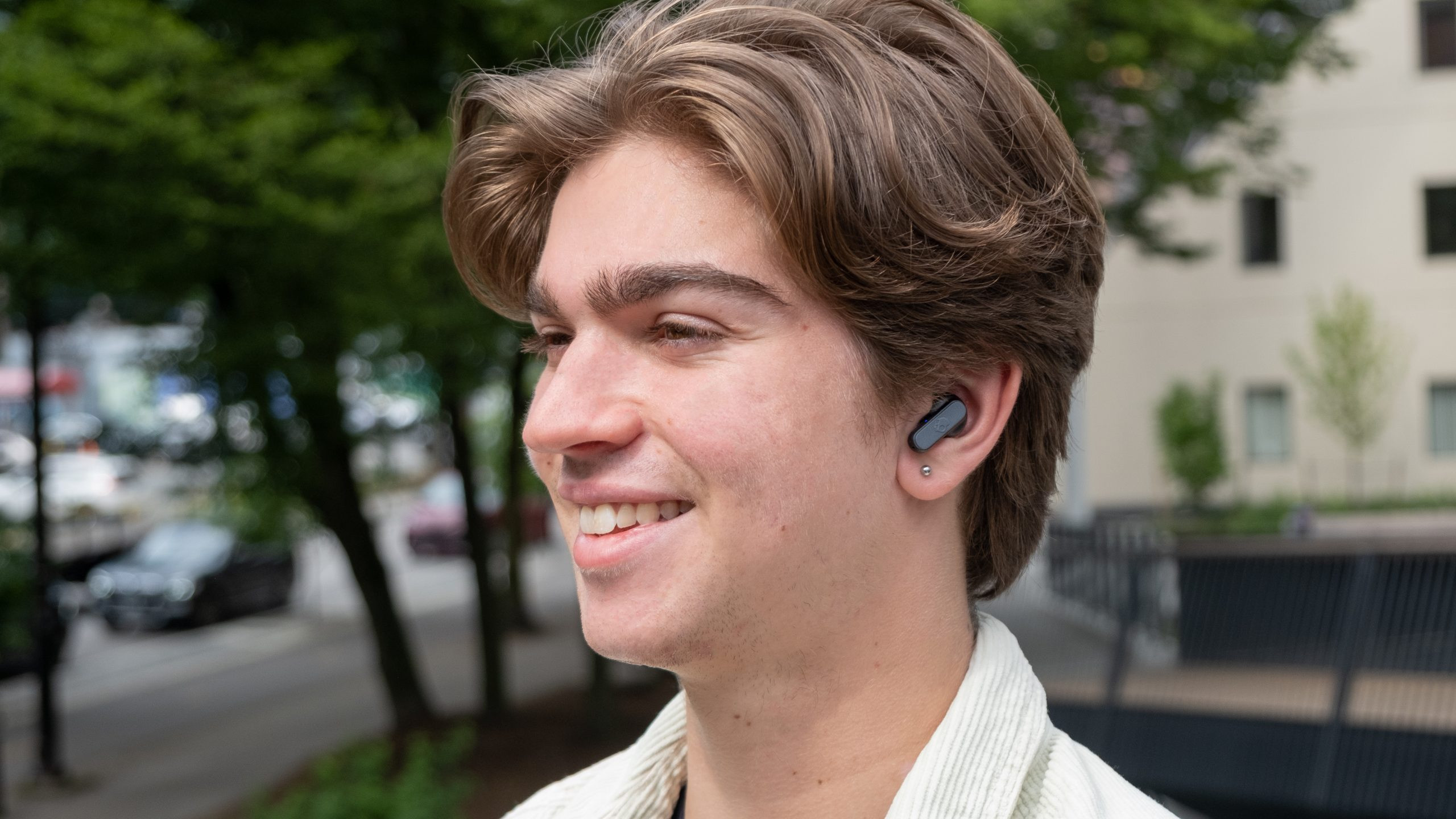 A man faces left smiling while wearing the Skullcandy Dime 2 with trees and a street in the background.