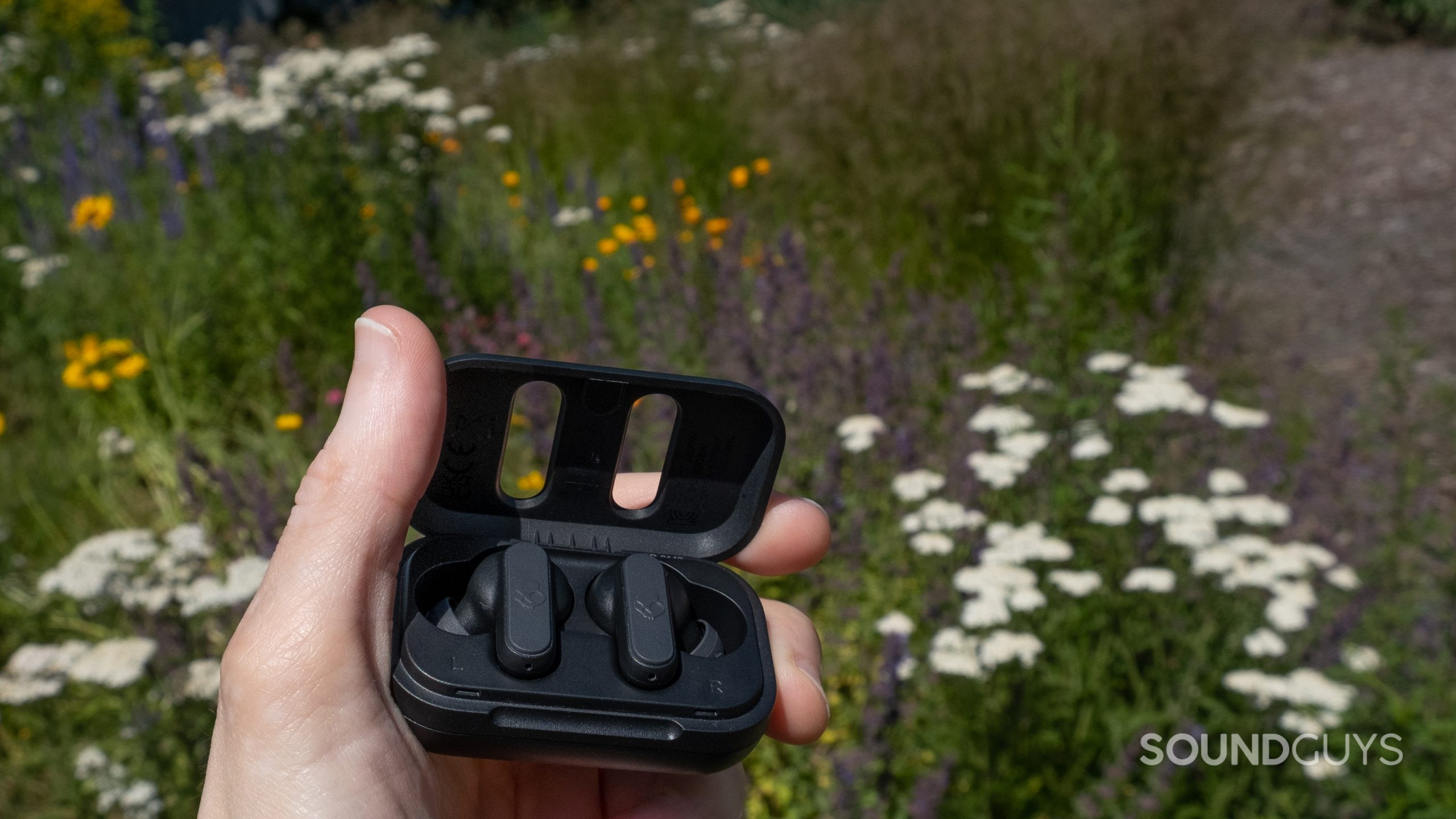 A hand holds the open case of the Skullcandy Dime 2 in front of a garden of wildflowers.