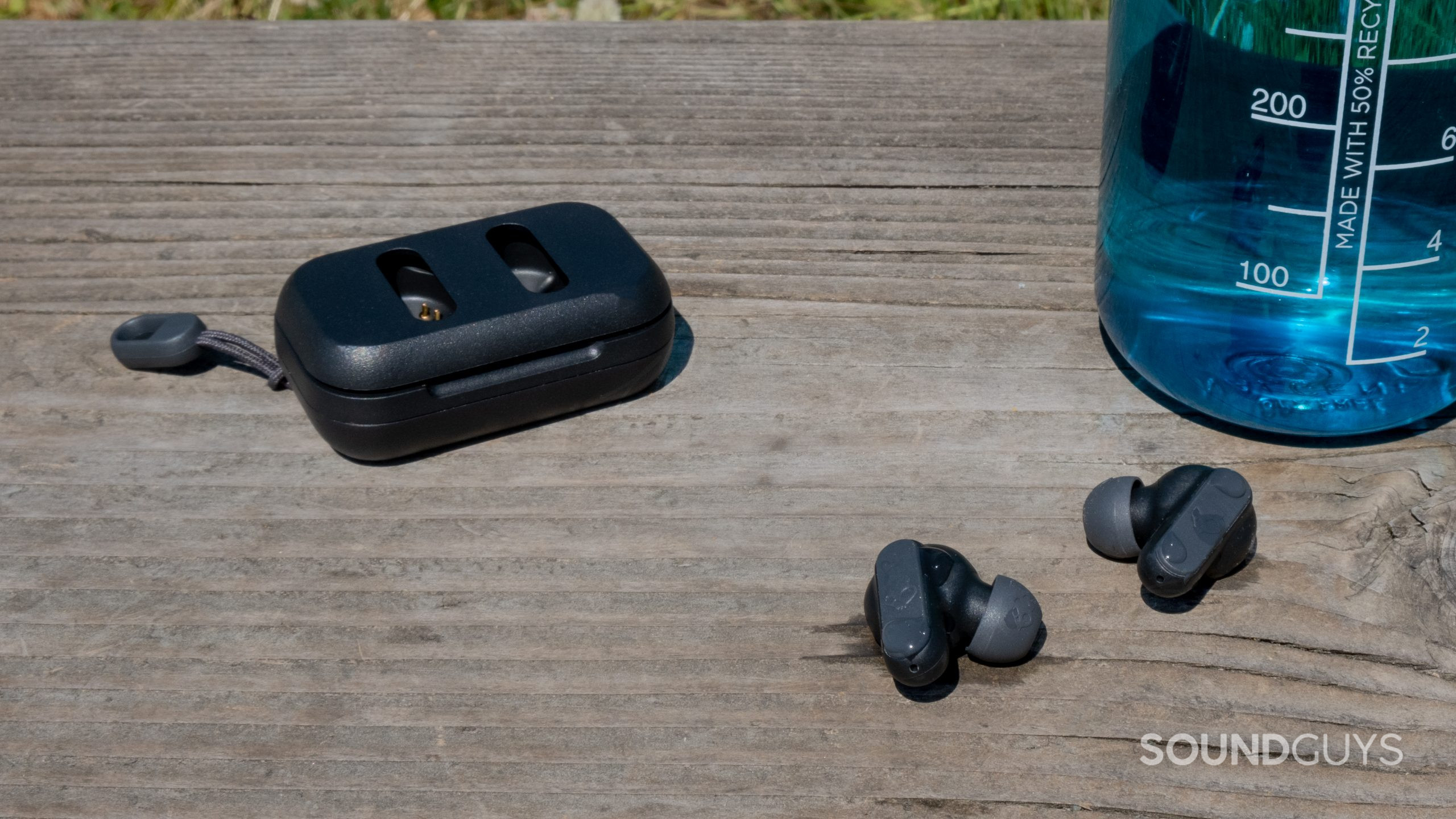 On a wood surface outside the Skullcandy Dime 2 buds have droplets of water on them next to a water bottle and the case.