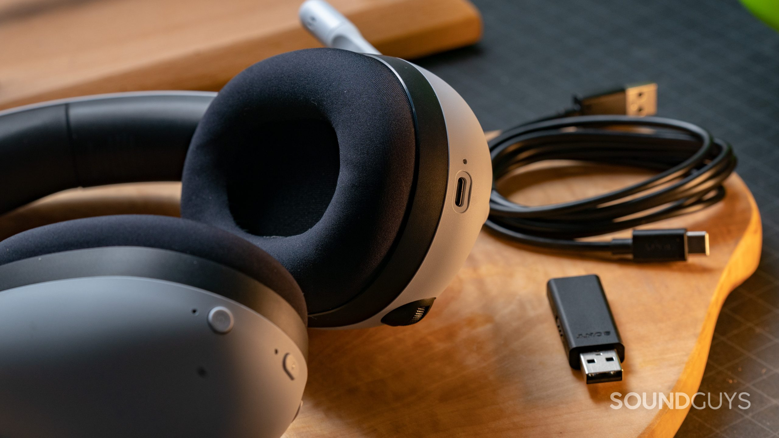 The Sony INZONE H7 gaming headset lays on wooden surface next to its dongle and charging cord, ear pads and charging port in view