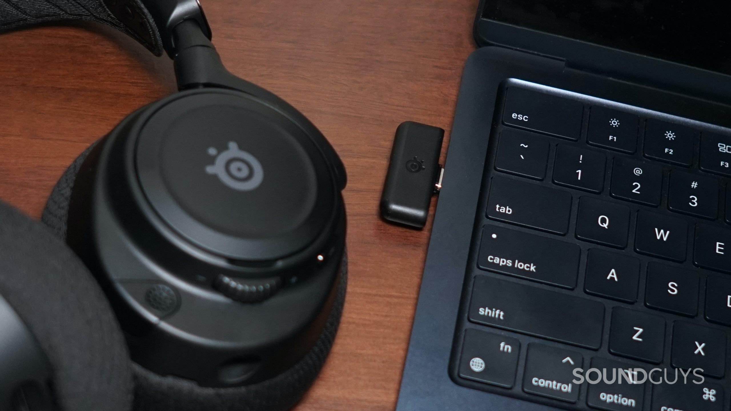 The SteelSeries Arctis Nova 7 lays on a wooden surface next to its USB-C dongle plugged into an Apple MacBook Air.