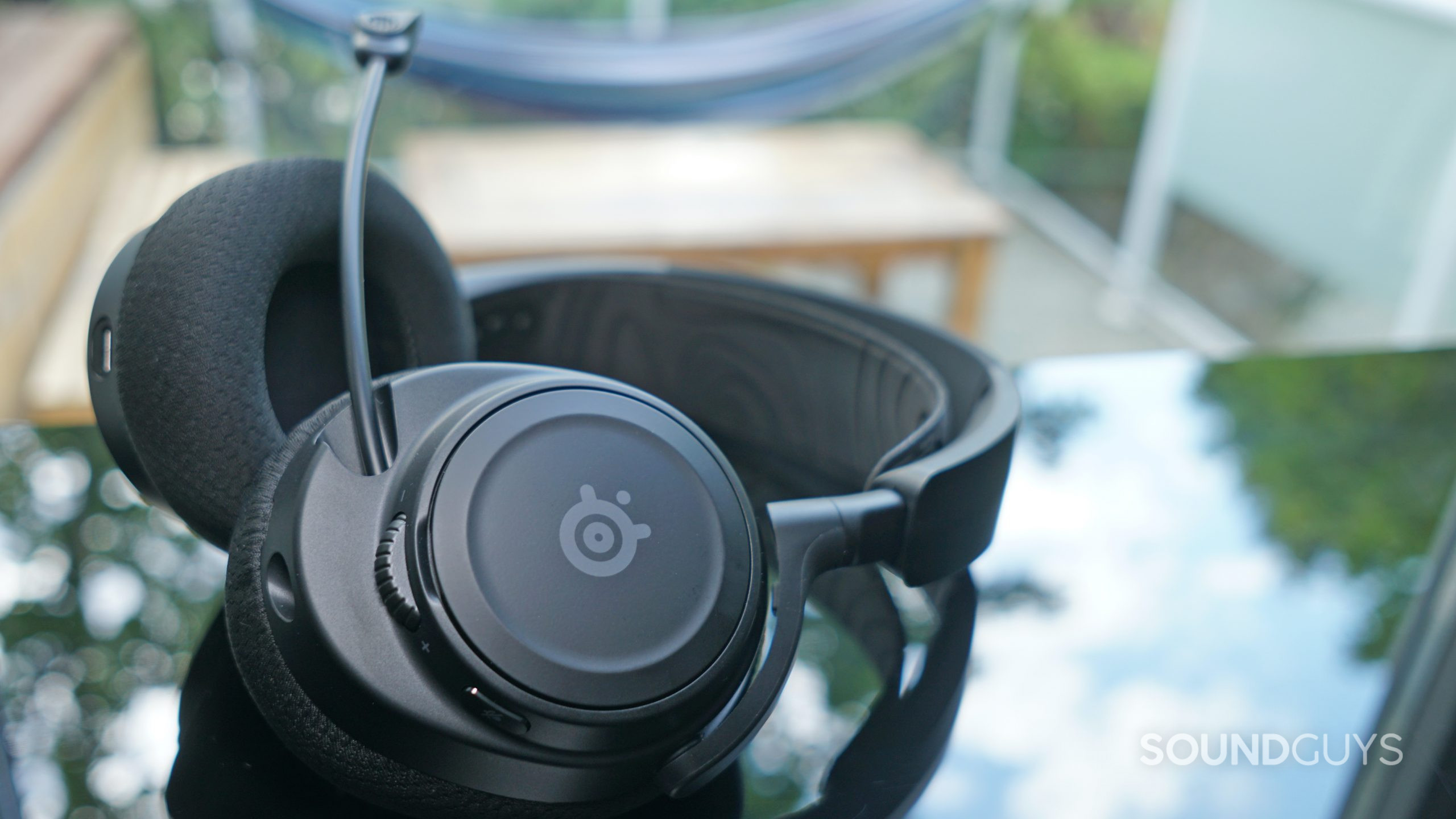 The SteelSeries Arctis Nova 7 lays on a reflective surface in front of a window