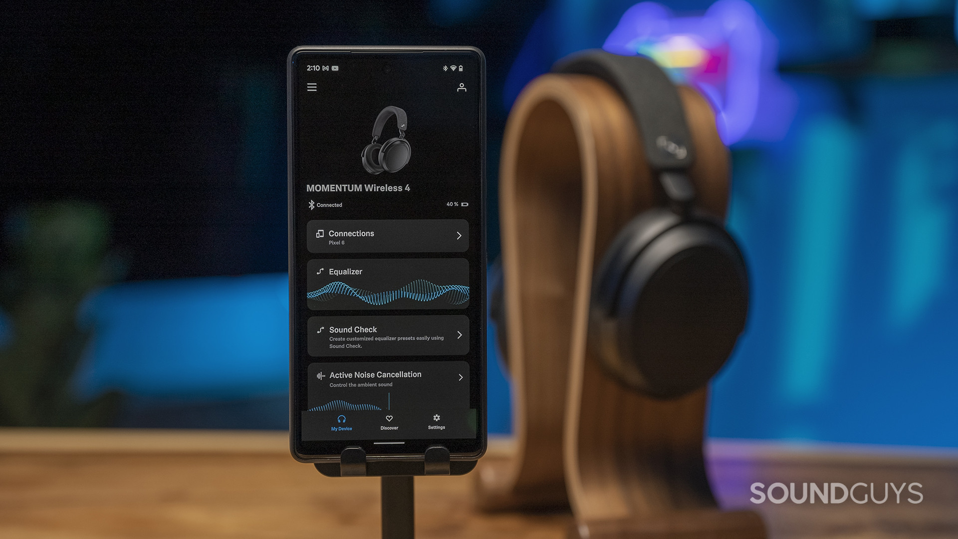 The Sennheiser Momentum 4 Wireless sits on a headphone stand in background with a phone displaying the Sennheiser Smart Control app displayed.