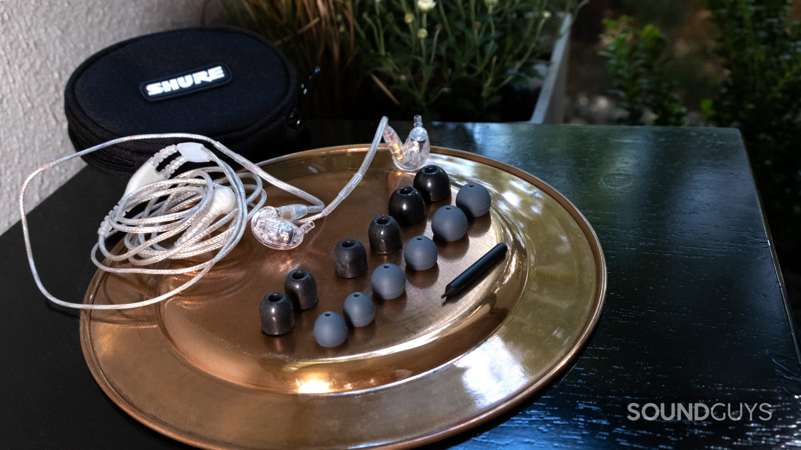 On a brass colored surface the full set of ear tips and the Shure SE215 sit outside on a black table with plants in the background