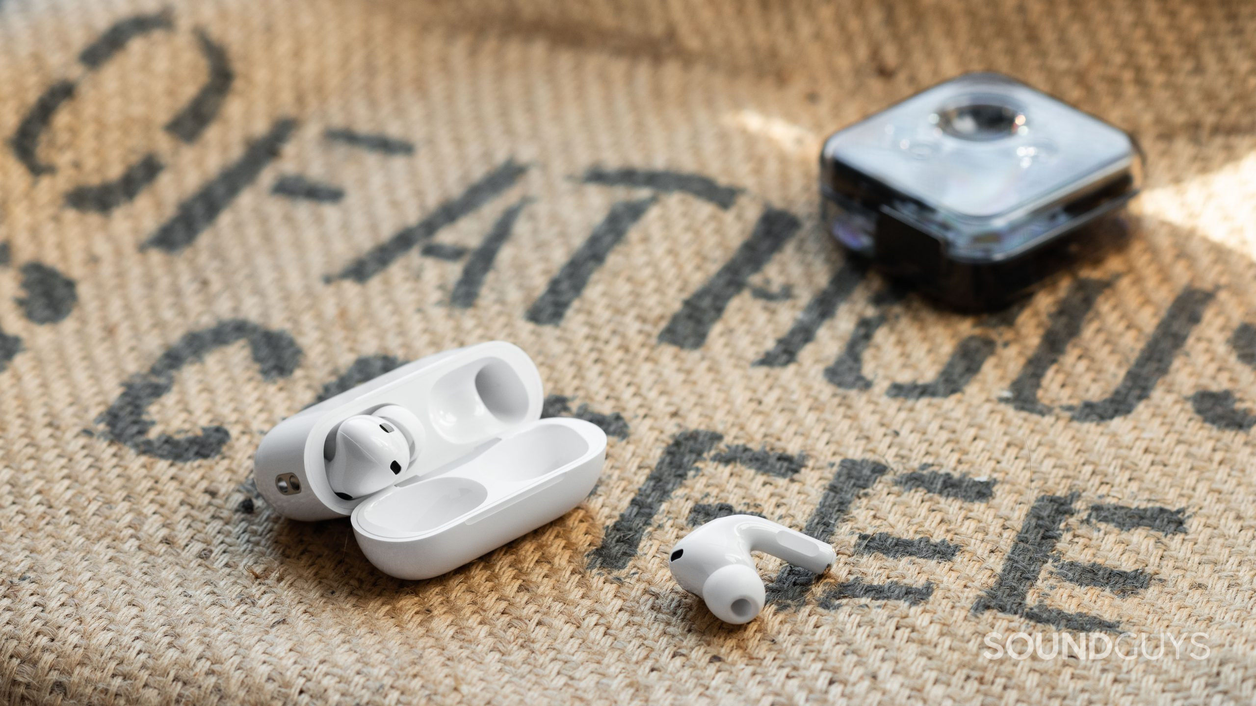 The Apple AirPods Pro (2nd generation) case open with one of the earbuds on a burlap sack, and the Nothing Ear 1 case in the background.