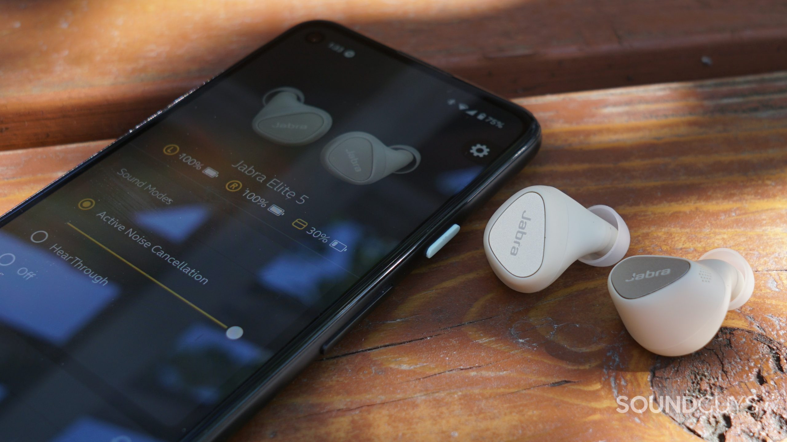 The Jabra Elite 5 earbuds lay on a wood surface next to a google Pixel 4a running the Jabra Sound+ app.