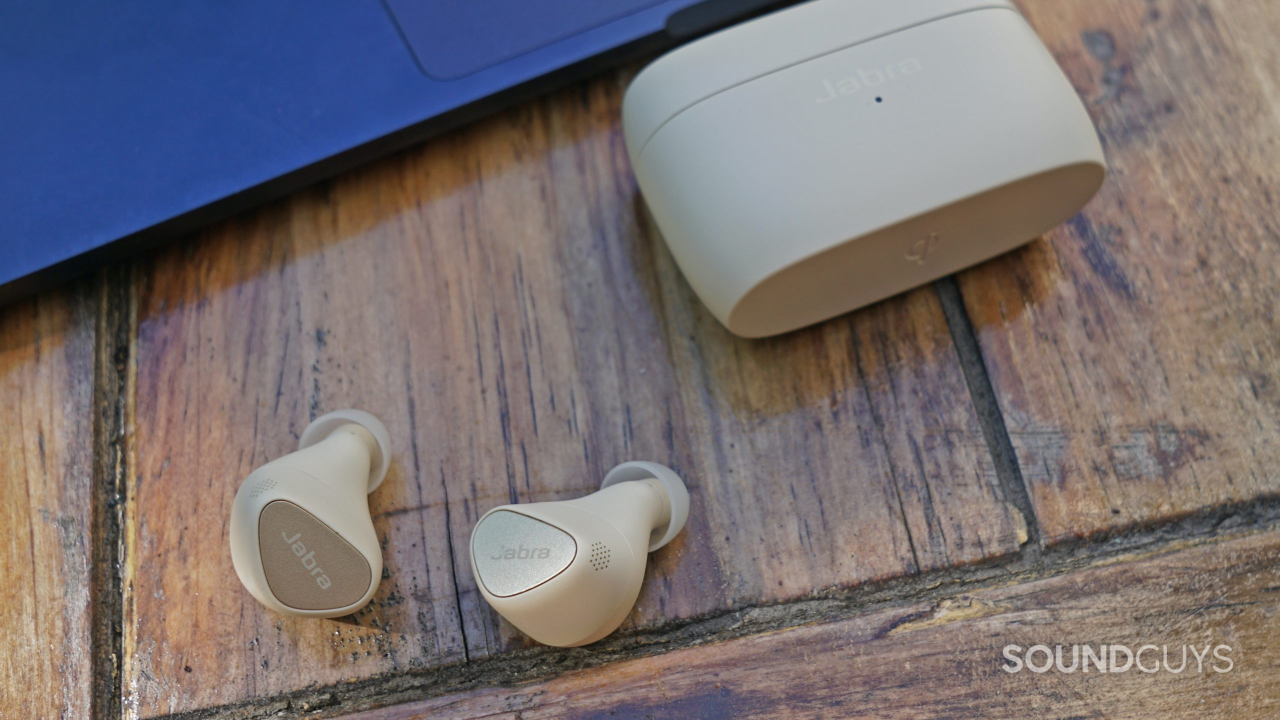 The Jabra Elite 5 true wireless earbuds lay on a wooden table next to its charging case and an Apple MacBook Air