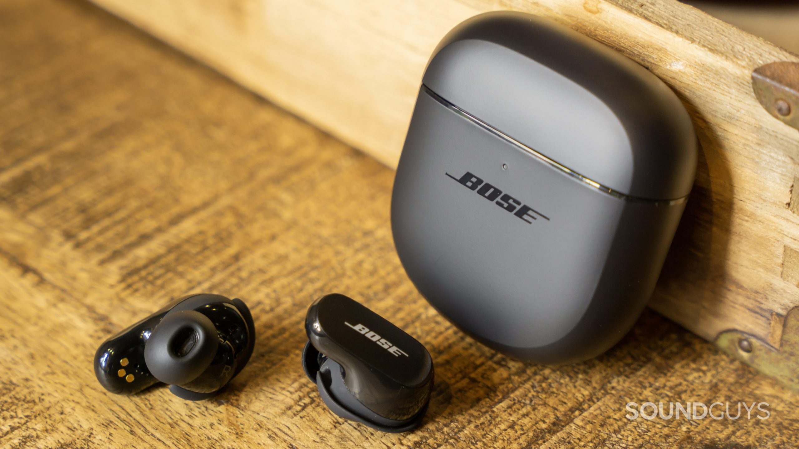 Propped against a wood surface the case of the Bose QuietComfort Earbuds II stands up with the buds out, showing the outside and inside faces of the housing.