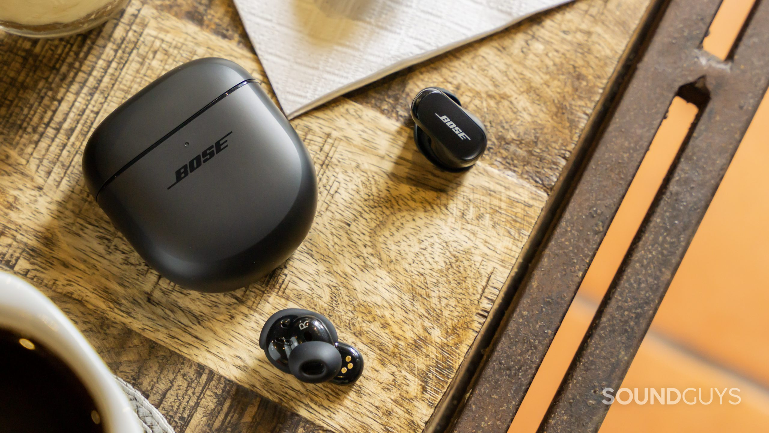 The Bose QuietComfort Earbuds II lays on a wooden table with coffee in the corner.