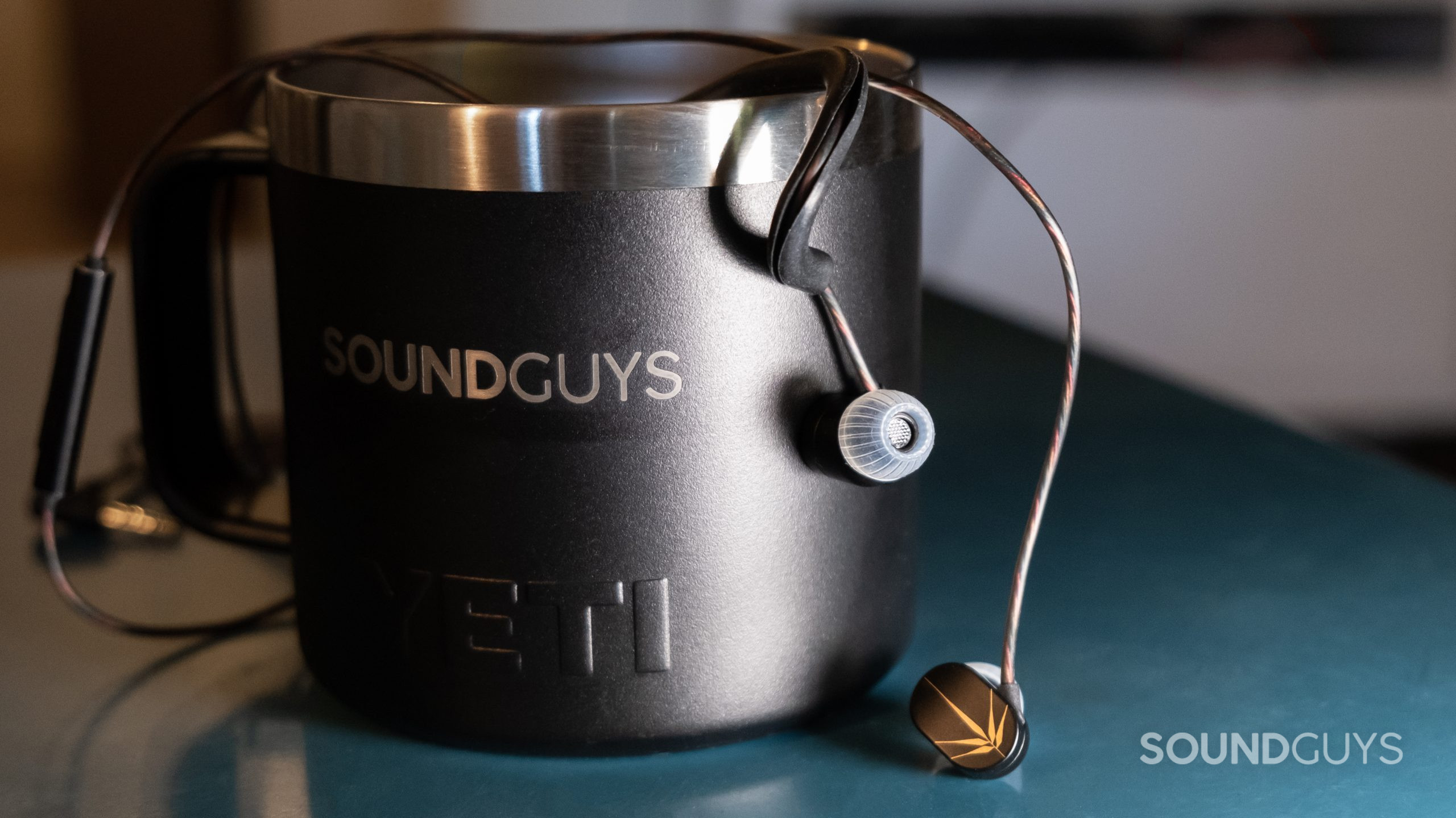 The Moondrop Chu hangs over the side of a SoundGuys branded mug on a teal metal table.