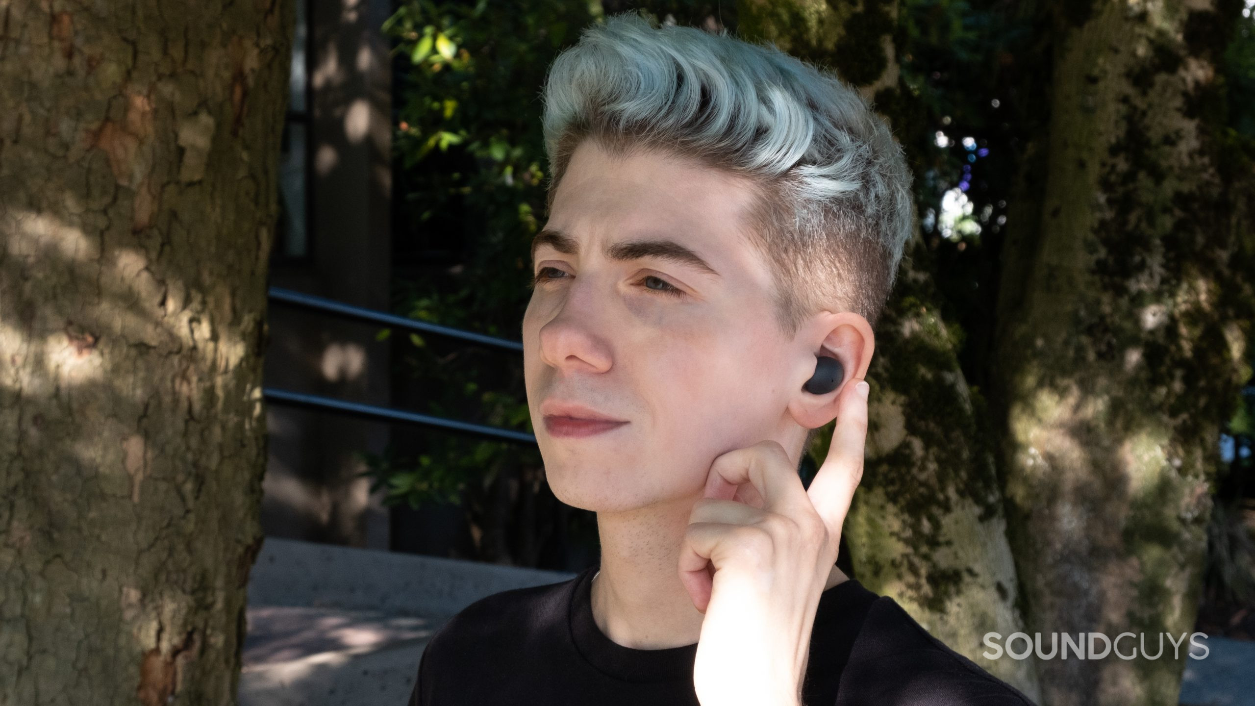 A man wears the Samsung Galaxy Buds 2 Pro while facing left, and he's about to tap the touch pad with trees and concrete in the background.