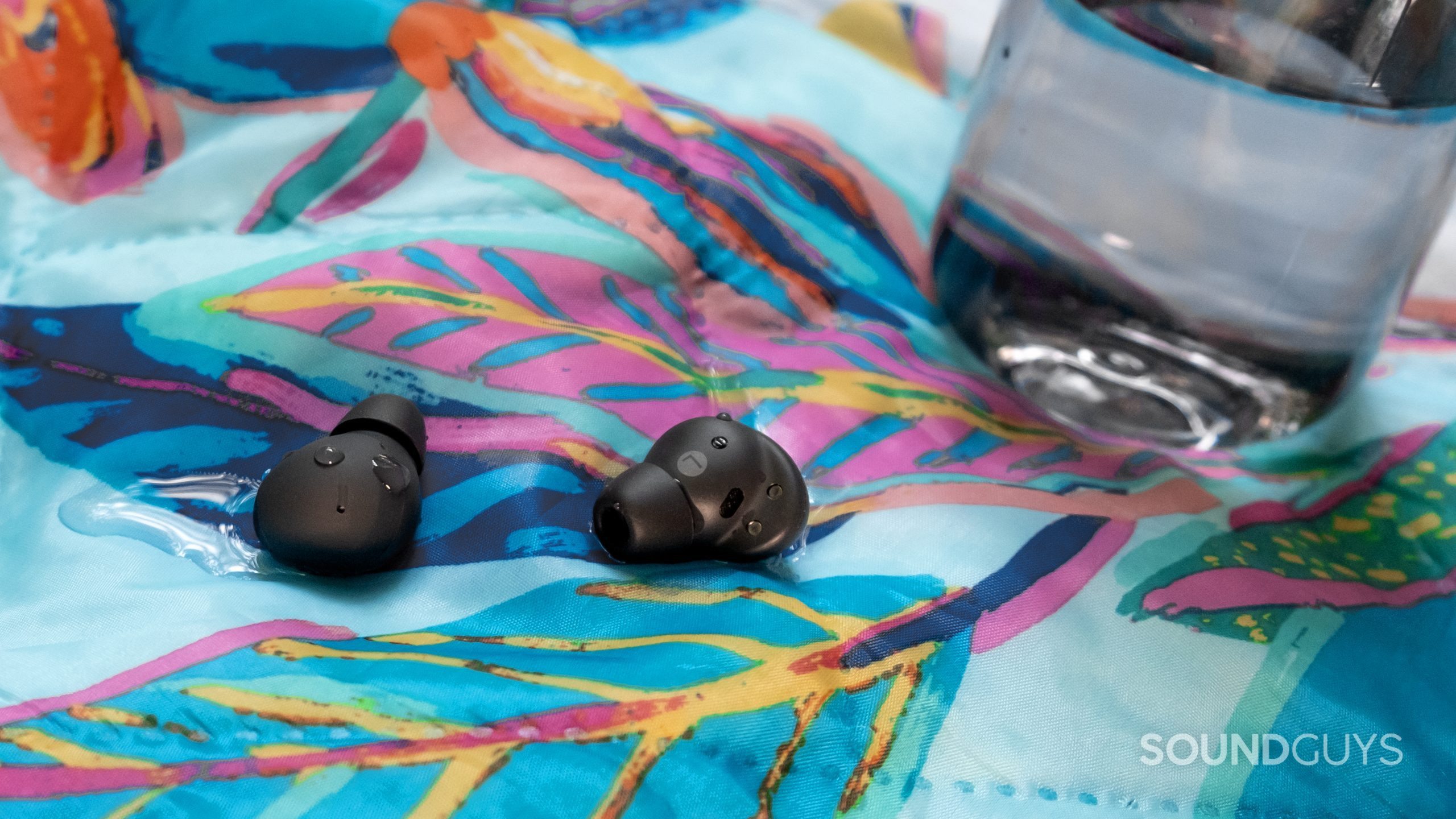 The Samsung Galaxy Buds 2 Pro are sitting in and covered in water droplets on a tropical themed outdoor blanket with a water bottle in the background.