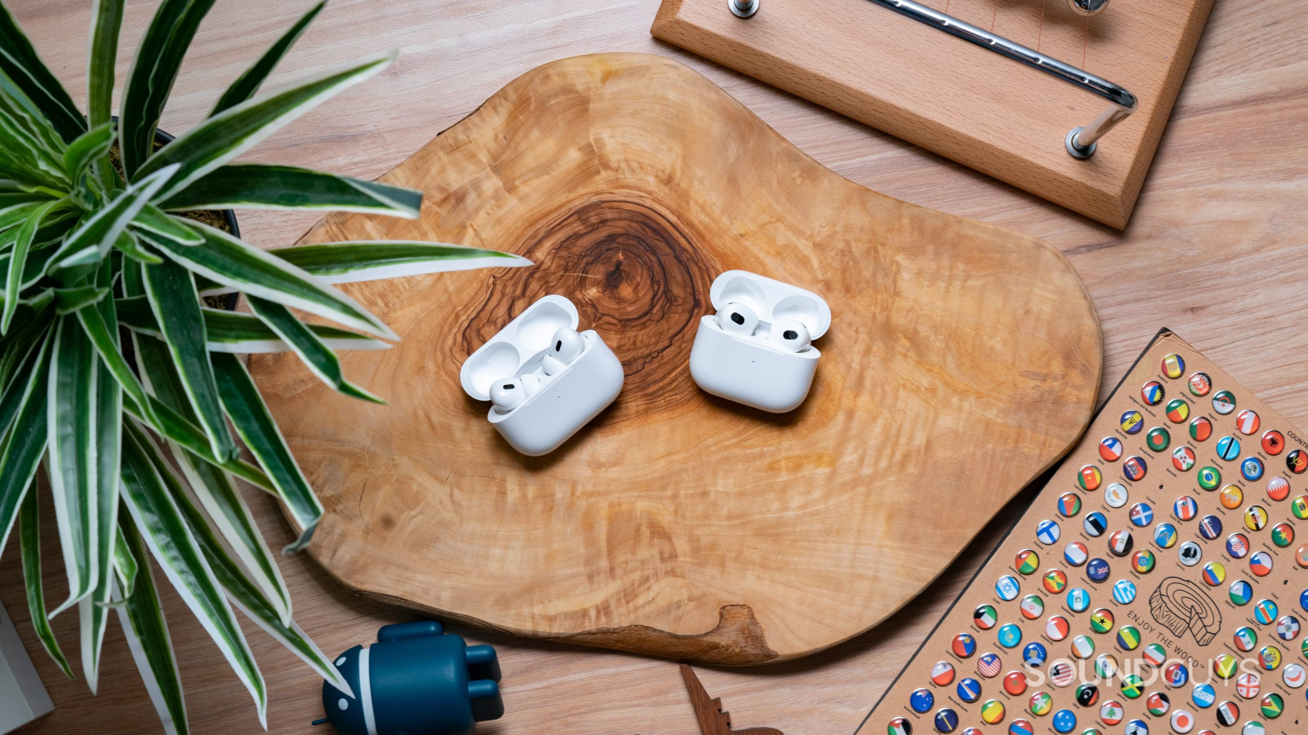 The Apple AirPods Pro Gen 2 and the Apple AirPods Gen 3 sitting across from each other with their cases open on top of a wooden surface.