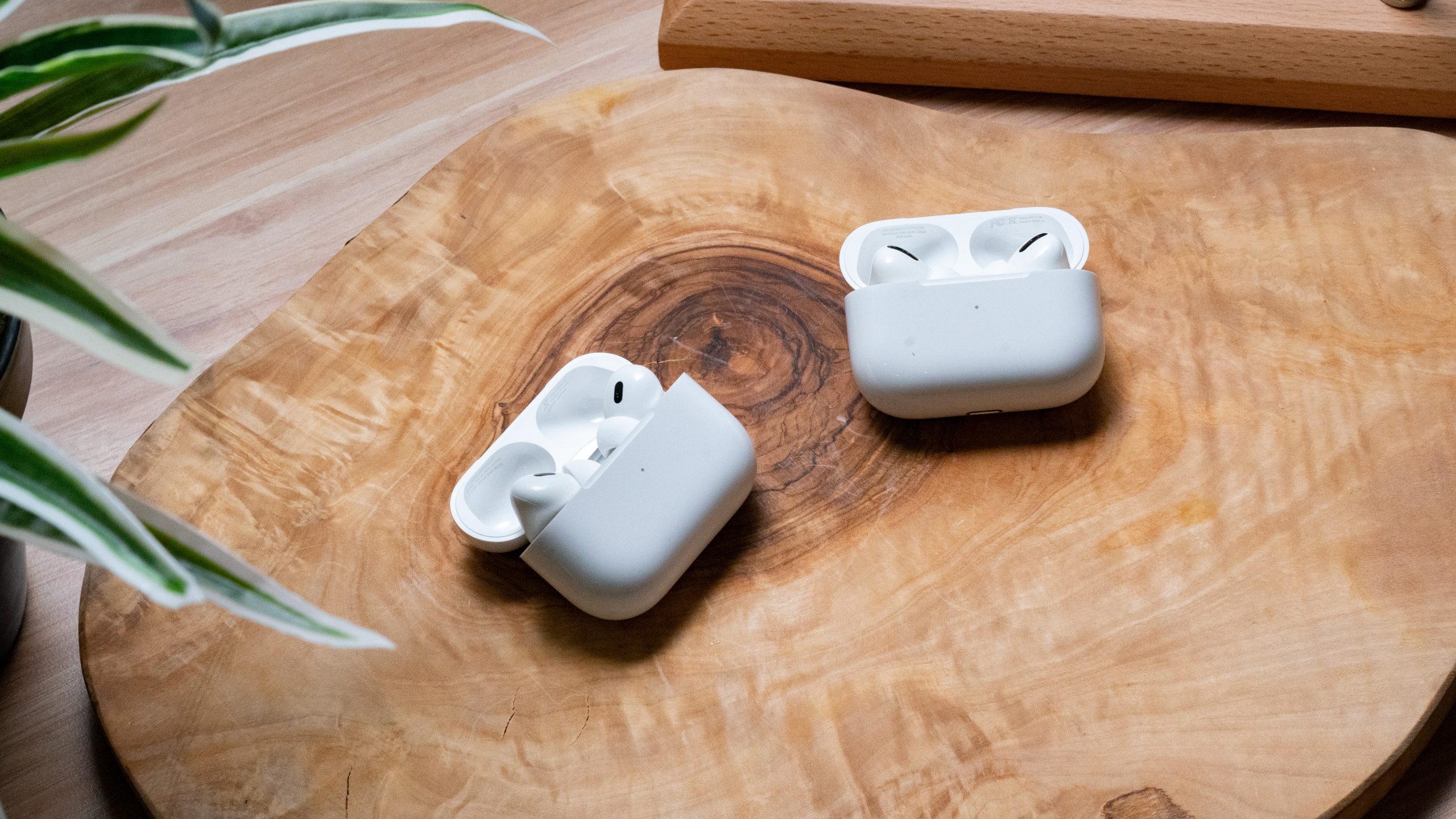 The Apple AirPods Pro (2nd generation) lays on a wooden surface next to the Apple AirPods Pro (1st generation), earbuds in open cases.