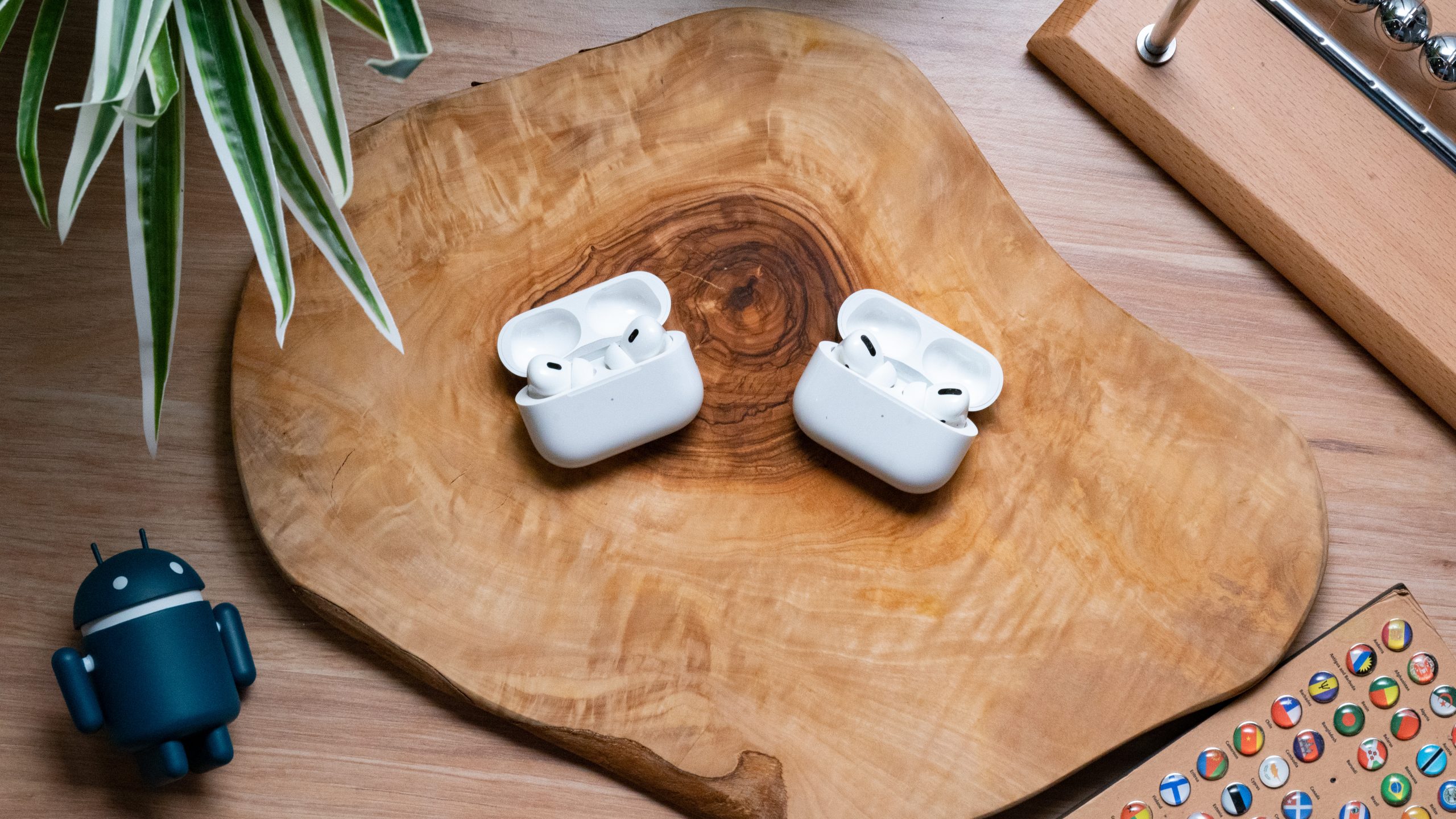 The Apple AirPods Pro (2nd generation) lays on a wooden surface next to the Apple AirPods Pro (1st generation), earbuds in open cases, shot from above.