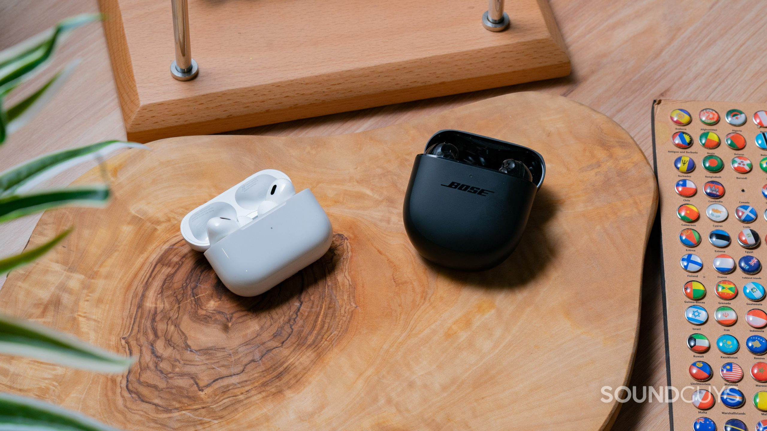 The AirPods Pro (2nd generation) and Bose QuietComfort Earbuds II in each open case beside each other on a wooden slab.