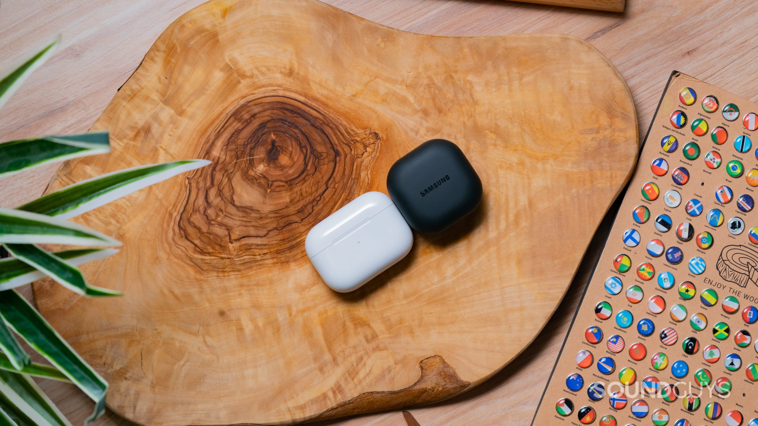 On a burled wood surface the Apple AirPods Pro (2nd generation) case rests on its back next to the Samsung Galaxy Buds 2 Pro charging case.