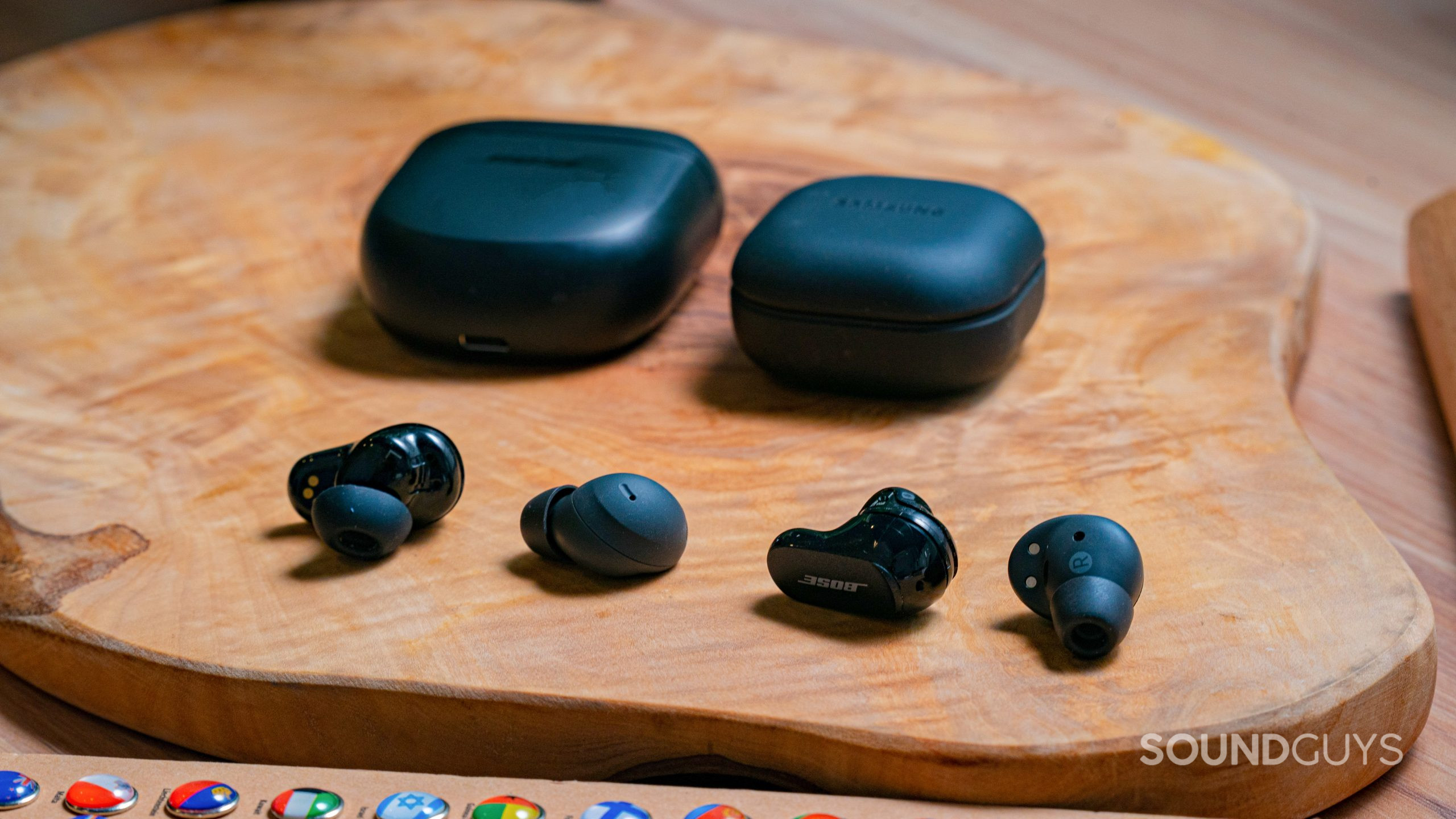The Bose QuietComfort Earbuds II and Samsung Galaxy Buds 2 Pro earbuds sitting side by side with the cases in the background on a wooden surface.