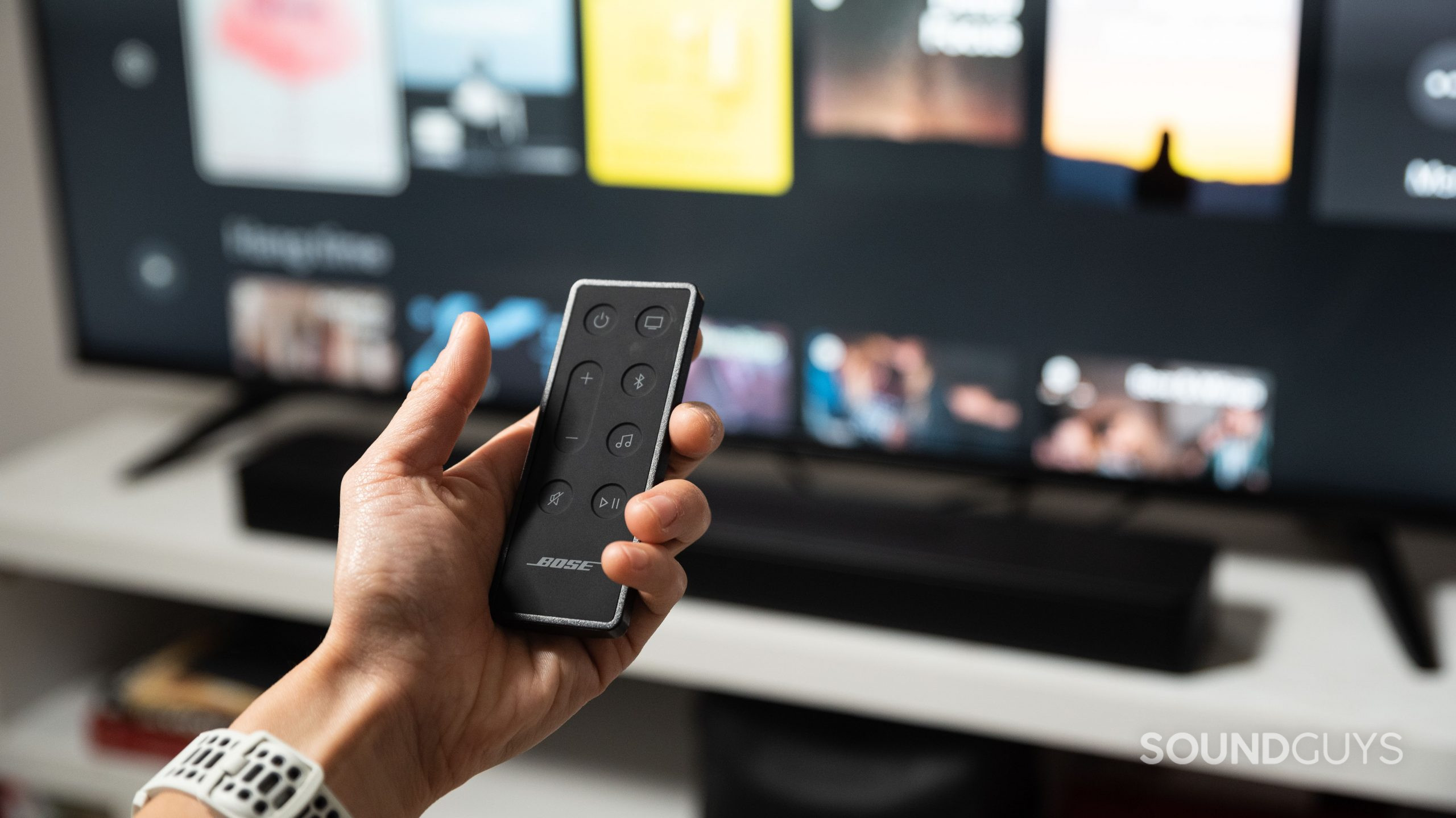 A hand holds the Bose Smart Soundbar 600 remote while pointing it at a TV.