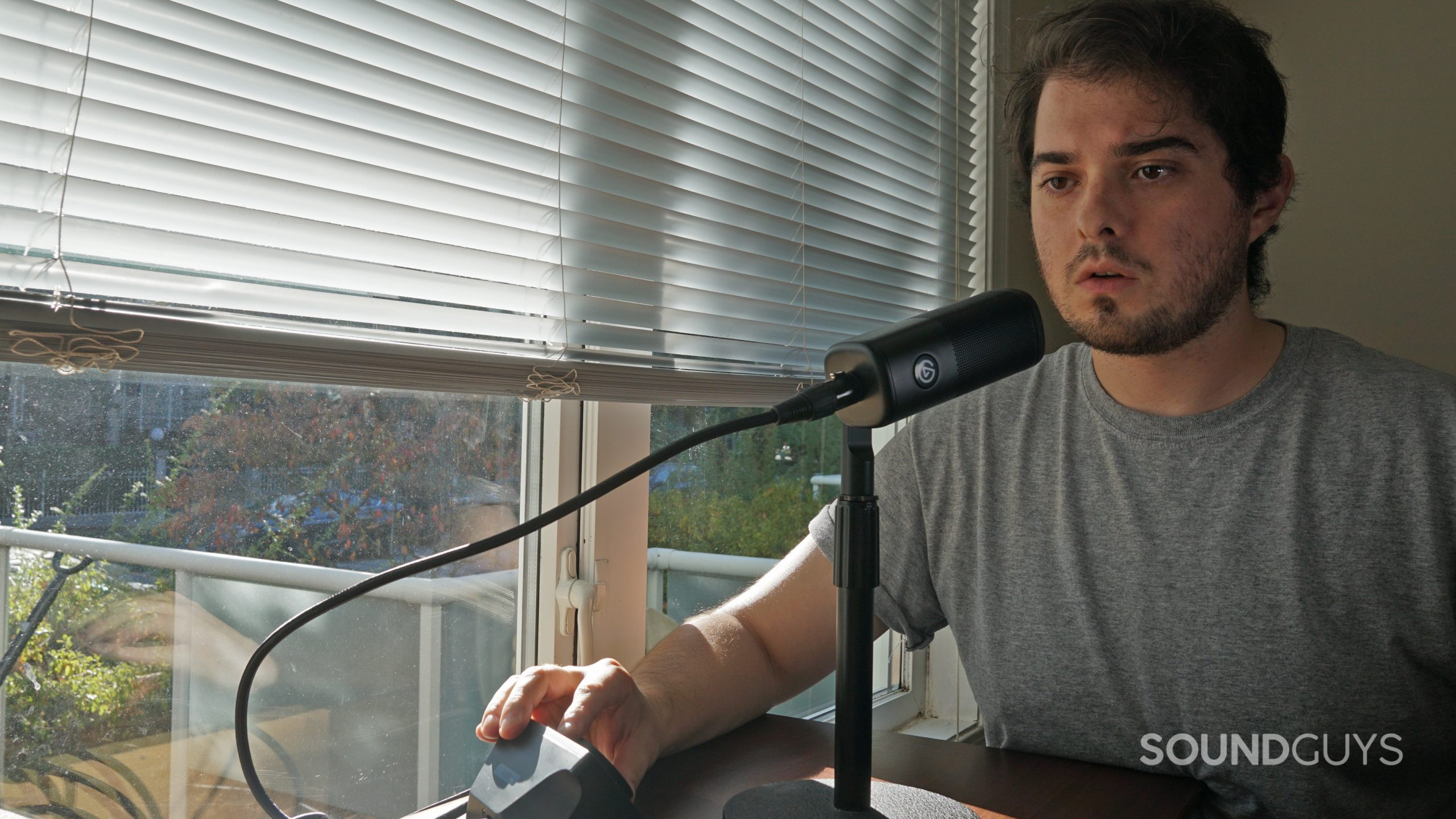A man stands next to a window and speaks into the Elgato Wave DX microphone, with a hand on the Elgato Wave DX.