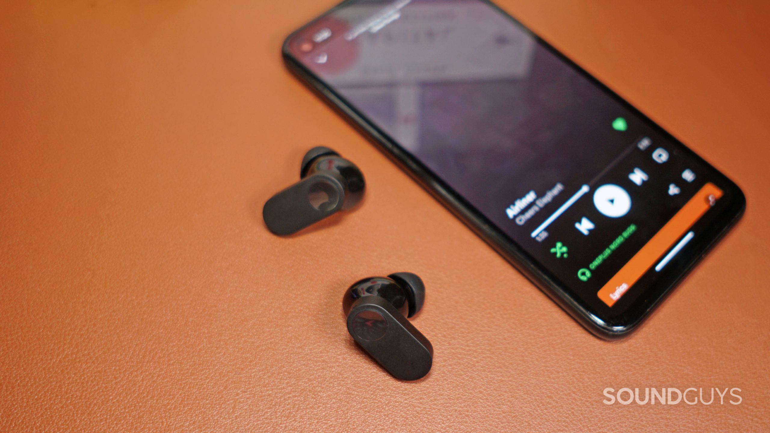 The OnePlus Nord Buds lays on a leather surface next to a Google Pixel 4a running spotify.