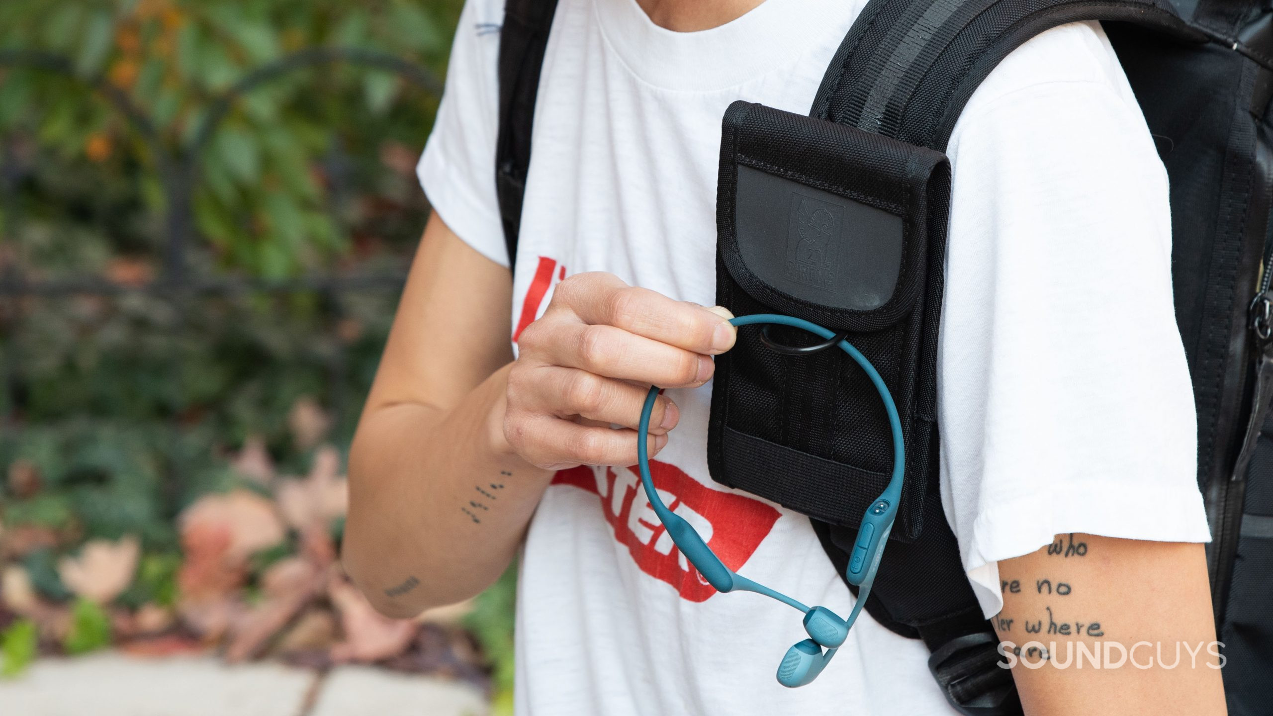 A person hooks the Shokz OpenRun Pro bone conduction headphones through a backpack phone pouch.