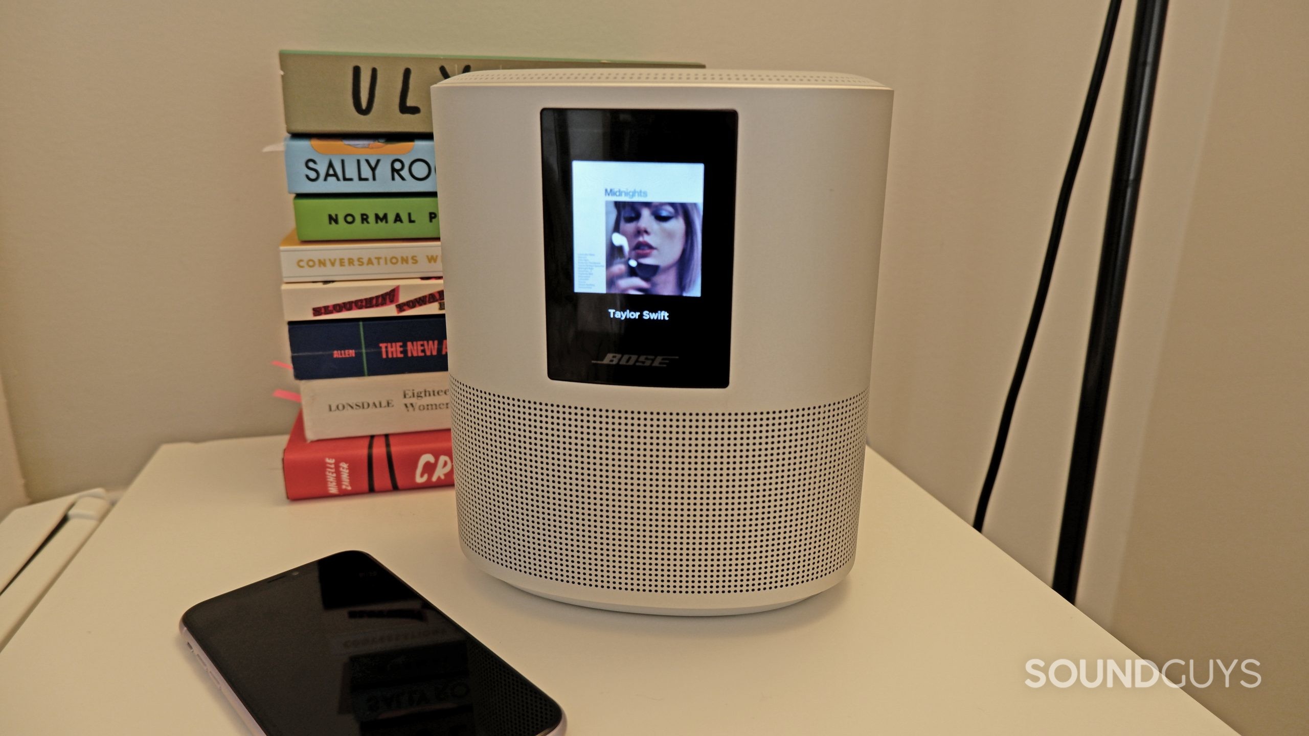 The Bose Home Speaker 500 sitting on an end table with a phone beside it, the speaker screen is showing that the speaker is playing Anti-Hero by Taylor Swift.