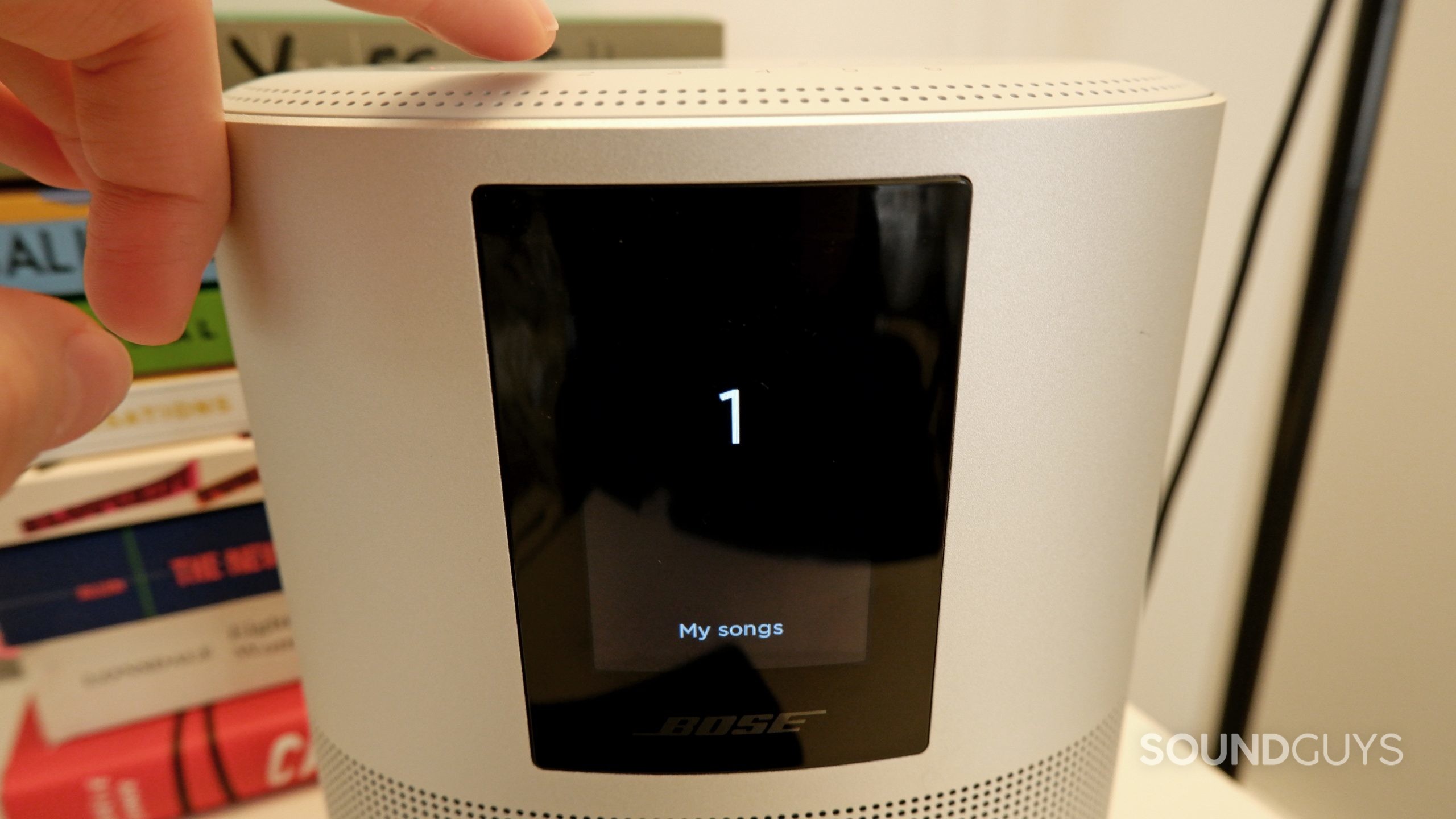 A hand pressing the first preset button on the Bose Home Speaker 500, with the screen showing the preset being chosen.