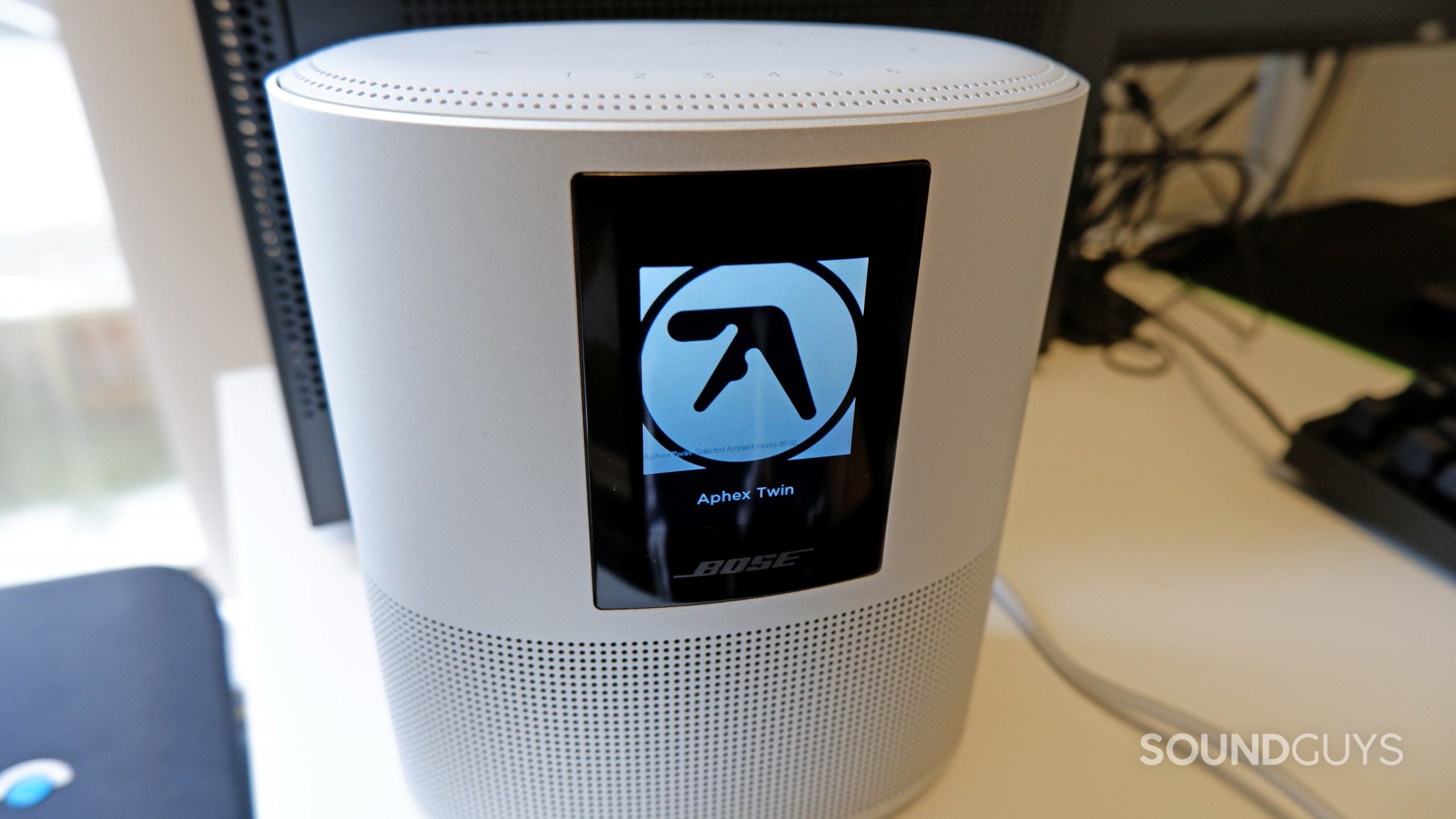 The Bose Home Speaker 500 playing Xtal by Aphex Twin, sitting on a white desk in front of a computer.