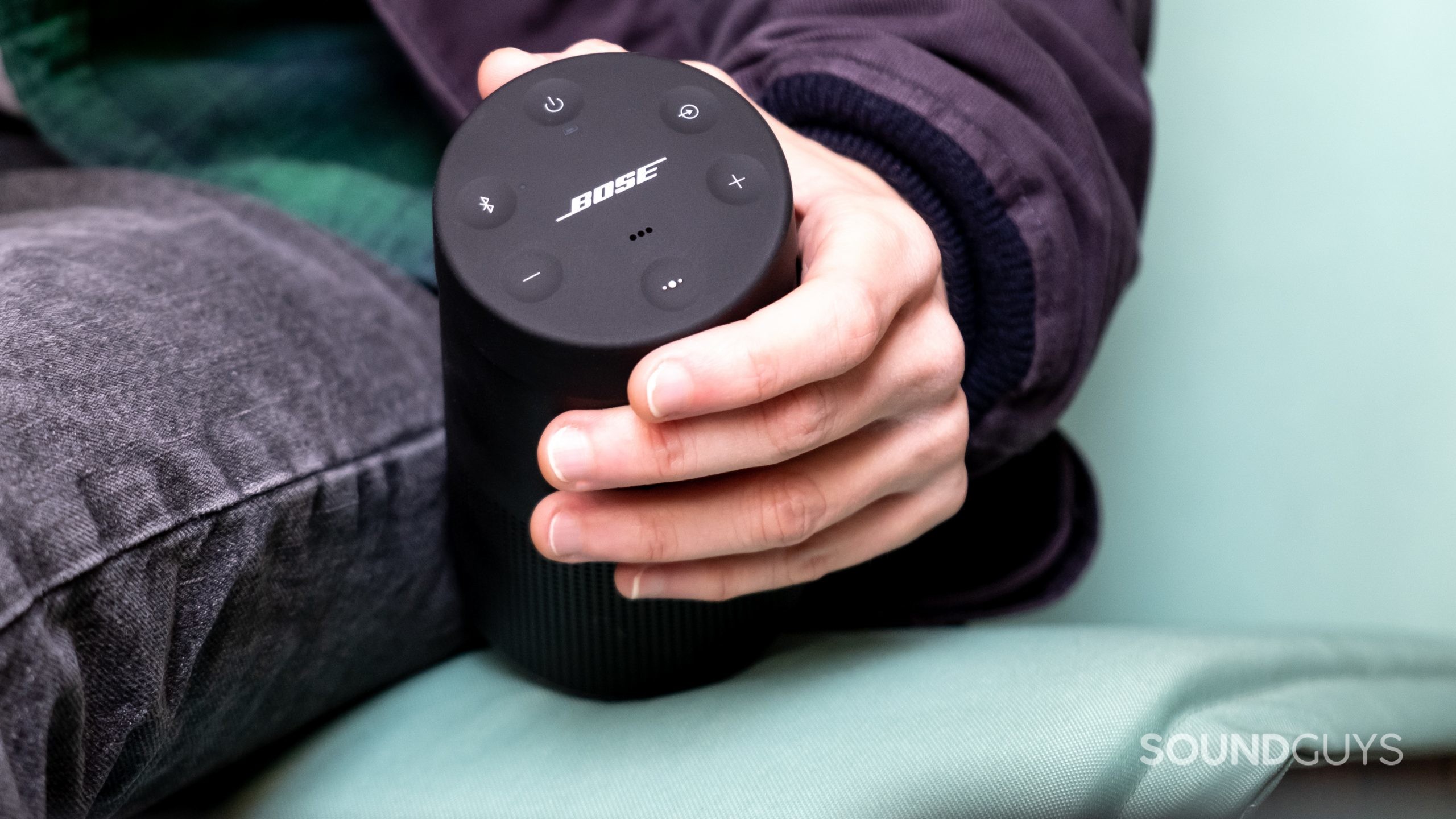 A person sits on a green cushion holding the Bose SoundLink Revolve II in hand.