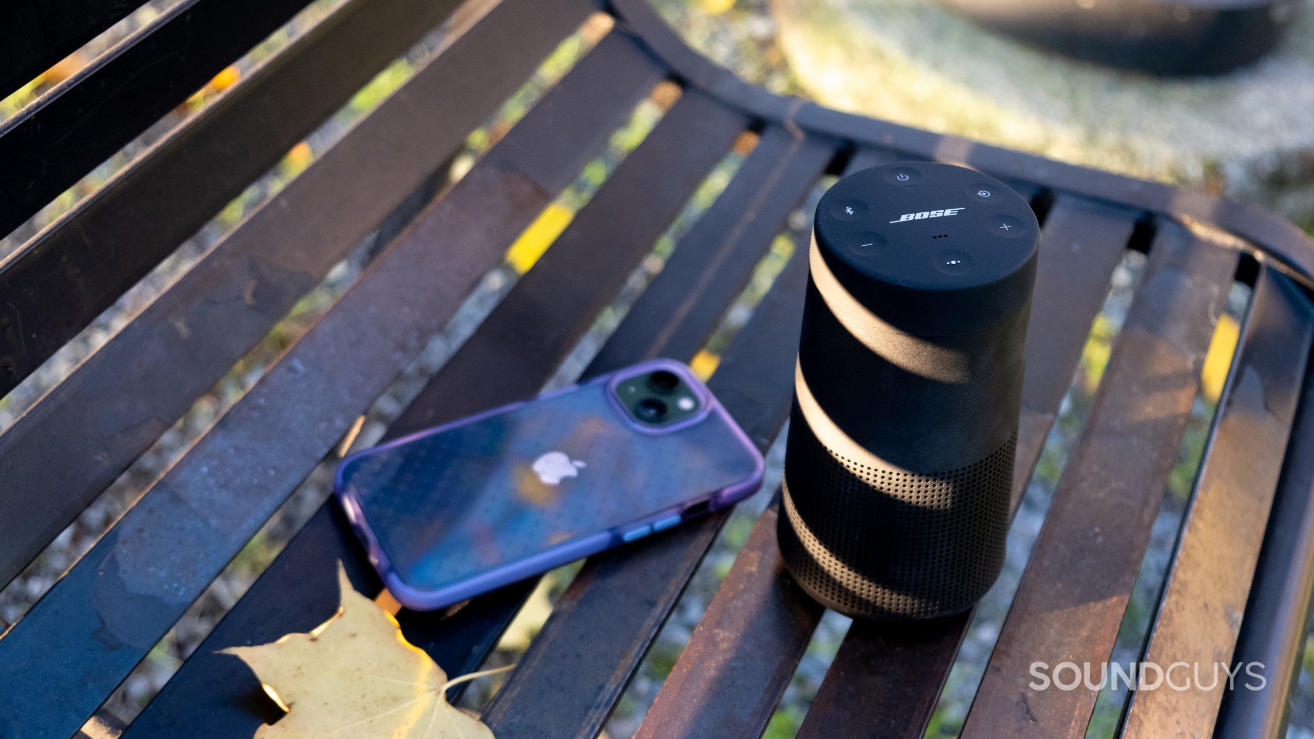 An iPhone sits on a metal bench next to a Bose SoundLink Revolve II with stripes of sunlight on the Bose speaker.