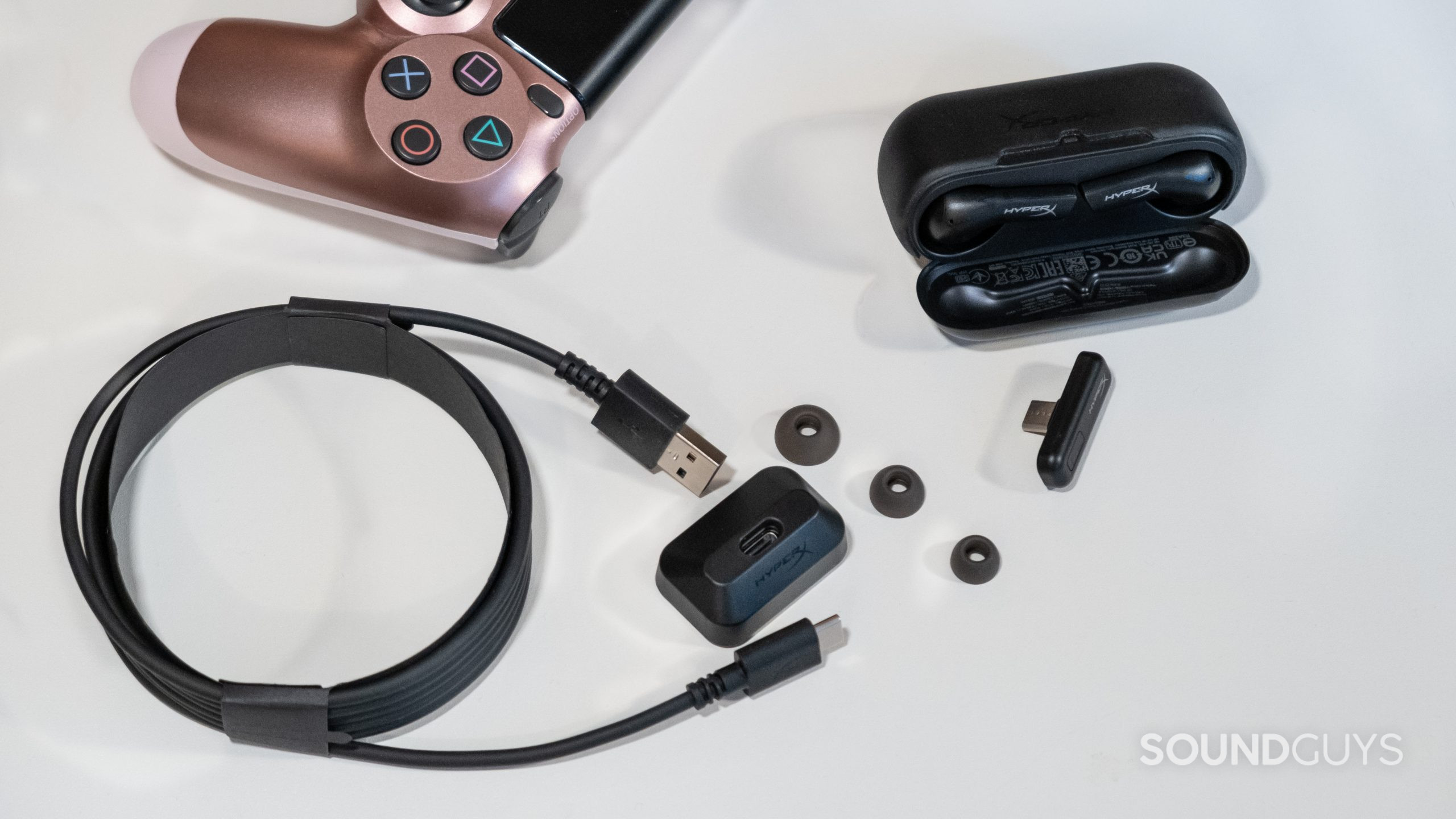 On an off white surface the open case of the HyperX Cloud Mix Buds with the included cable, USB-C dongle, ear tips, and a console controller are spread out.