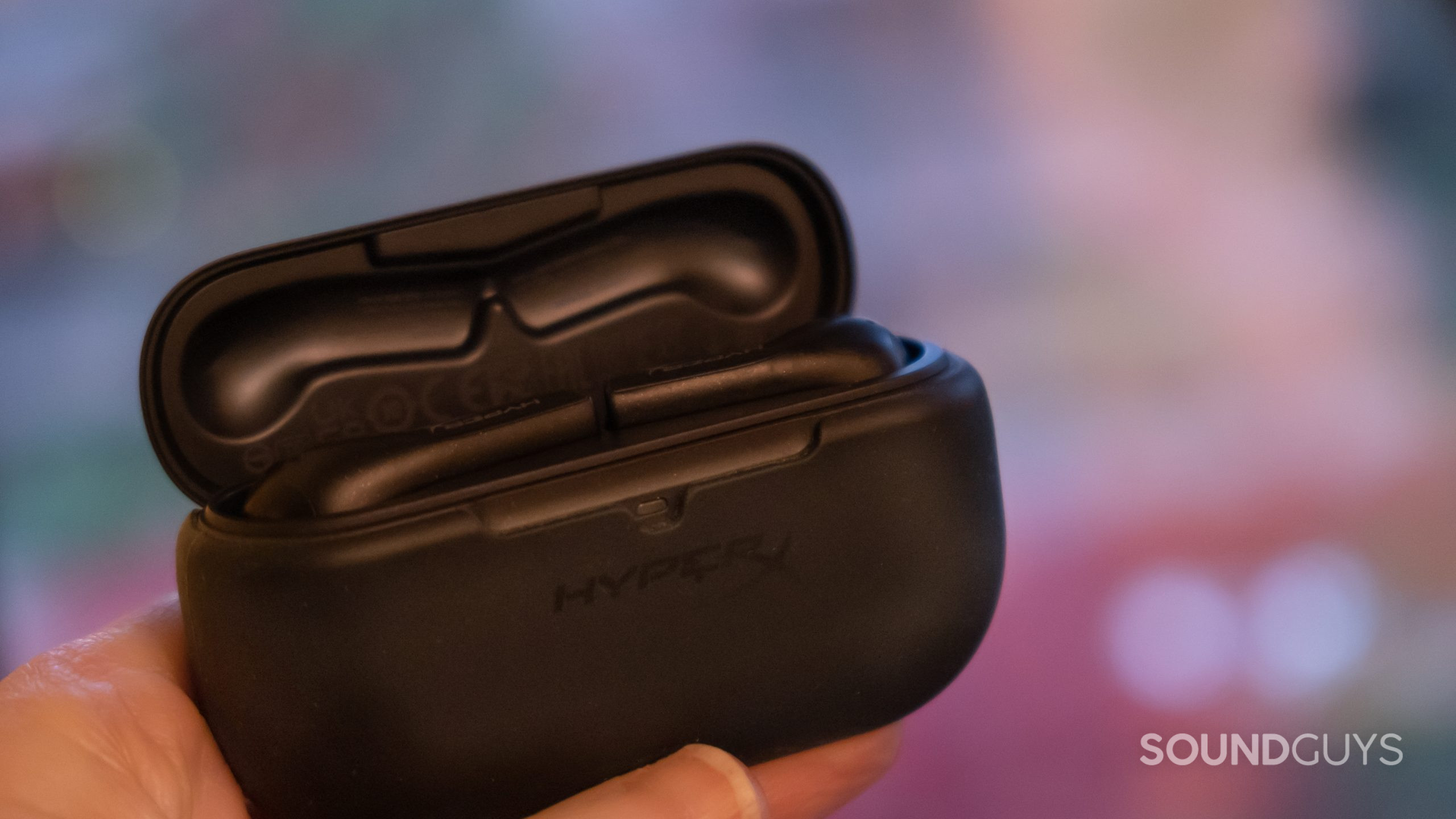 A hand holds up the front face of the open case of the HyperX Cloud Mix Buds with a blurred autumn background.