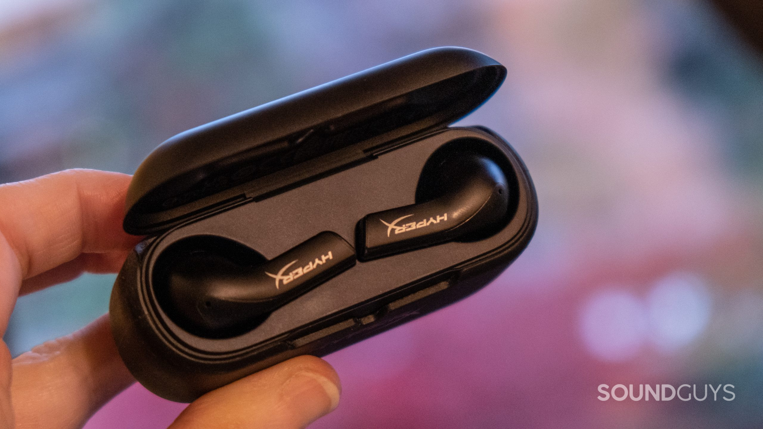 A hand holds the open case of the HyperX Cloud Mix Buds showing the tops of the buds set against a blurred autumn scene.