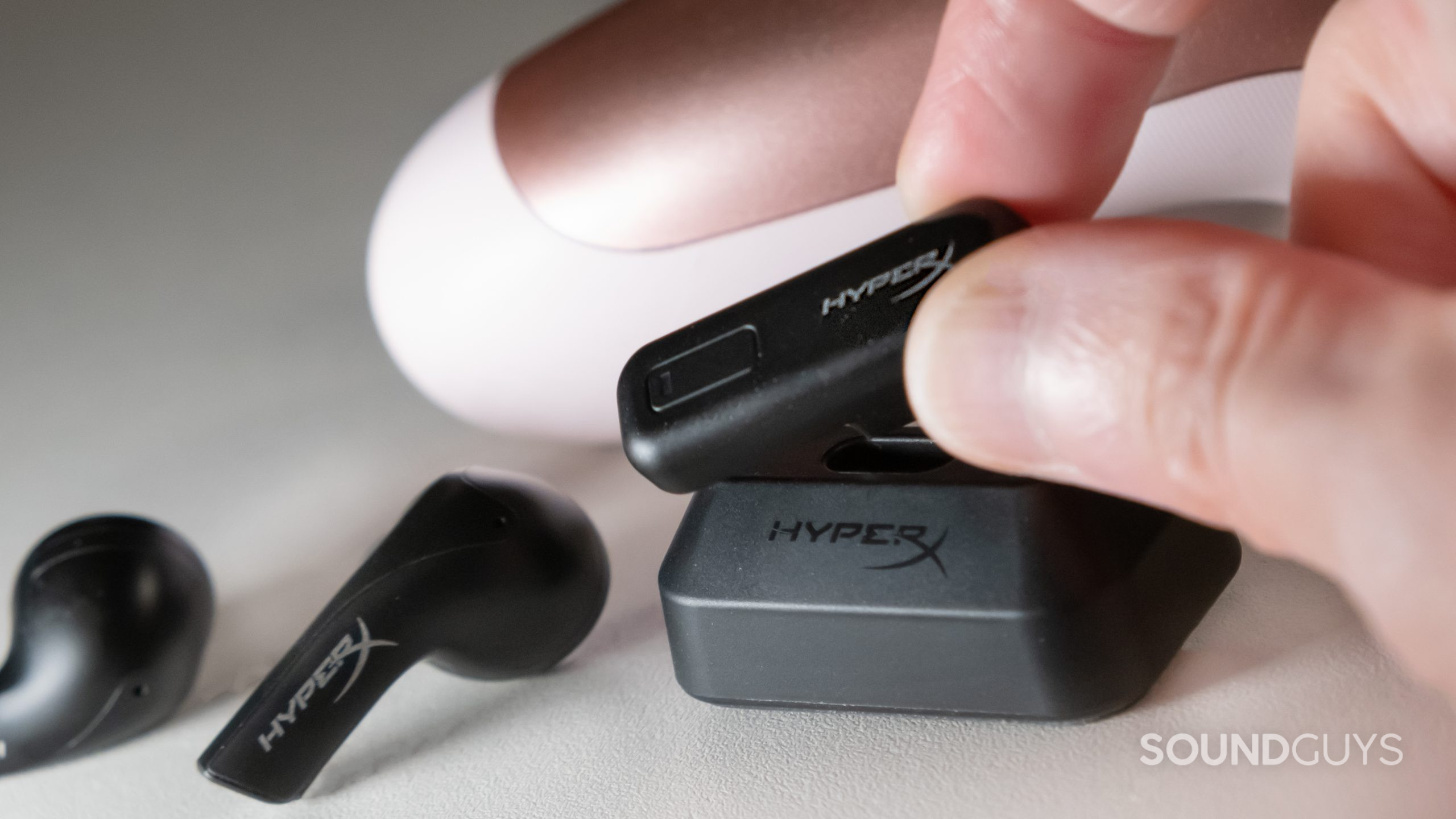 A hand is in the process of inserting the HyperX Cloud Mix Buds 2.4GHz USB-C dongle into the PC platform with the buds on the left and a controller in the background.