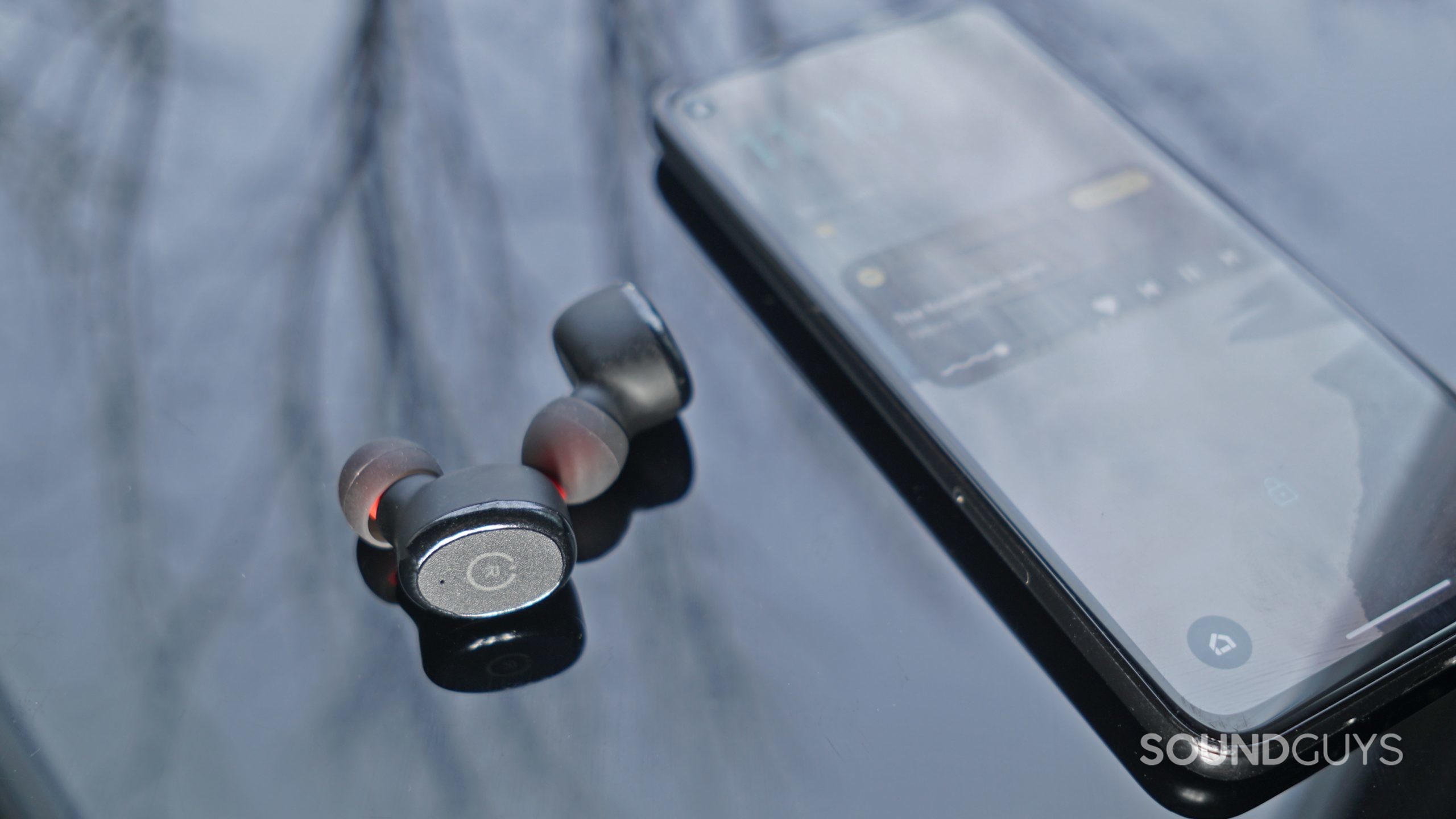 The TOZO T10 lays on a reflective black surface next to a Pixel 4a running Spotify