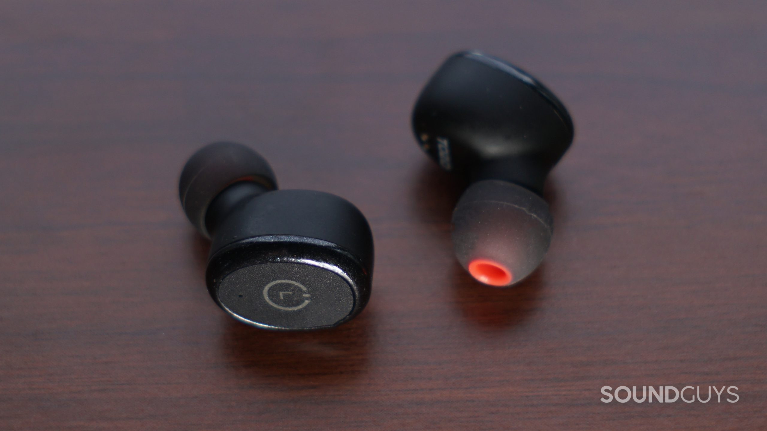 TOZO-T10-earbuds-on-wood The TOZO T10 lays on a wooden surface with the left earbud touch panel in view.