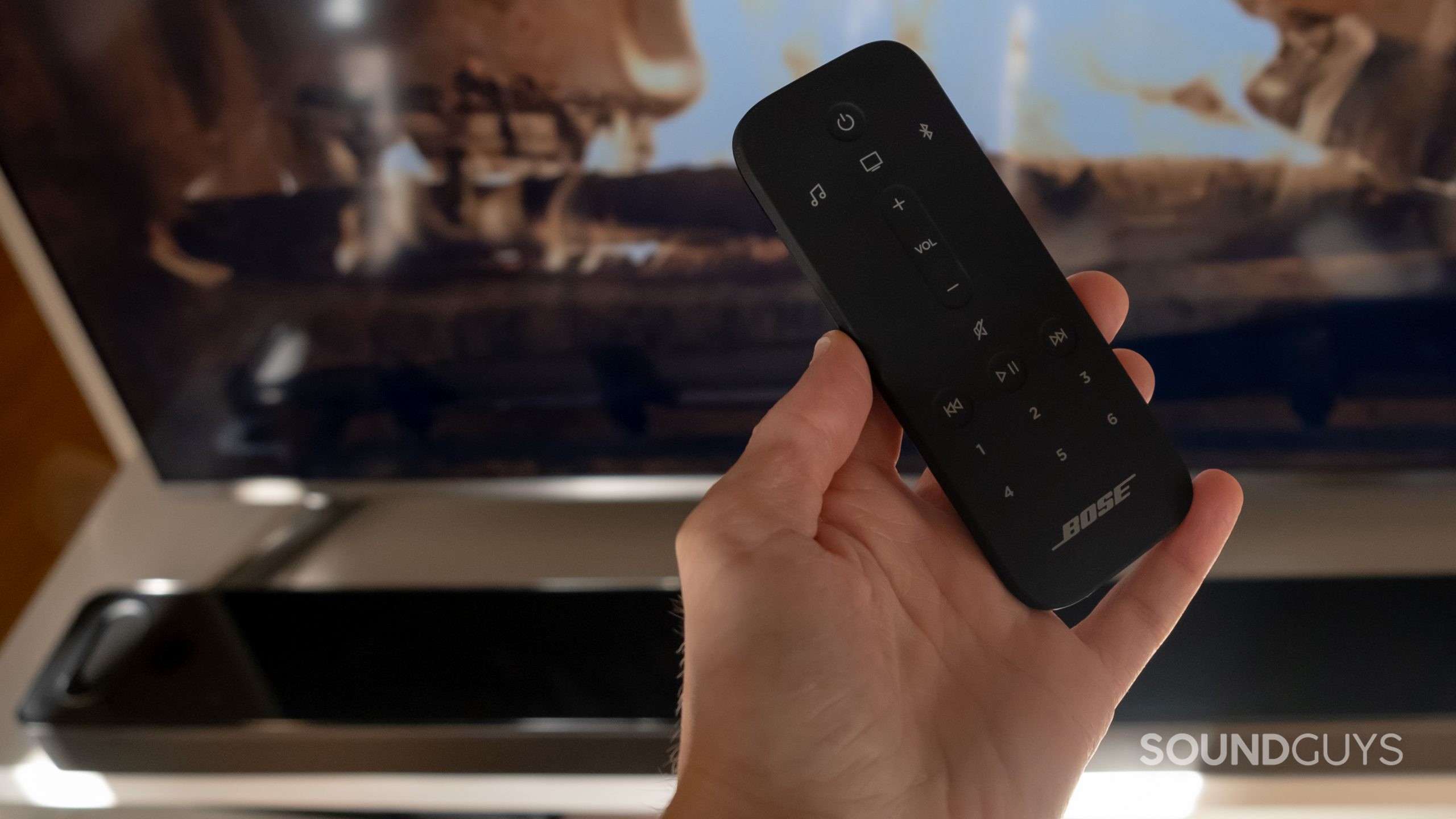 A hand holds the remote that comes with the Bose Smart Soundbar 900 in front of a TV and the soundbar itself in the background.
