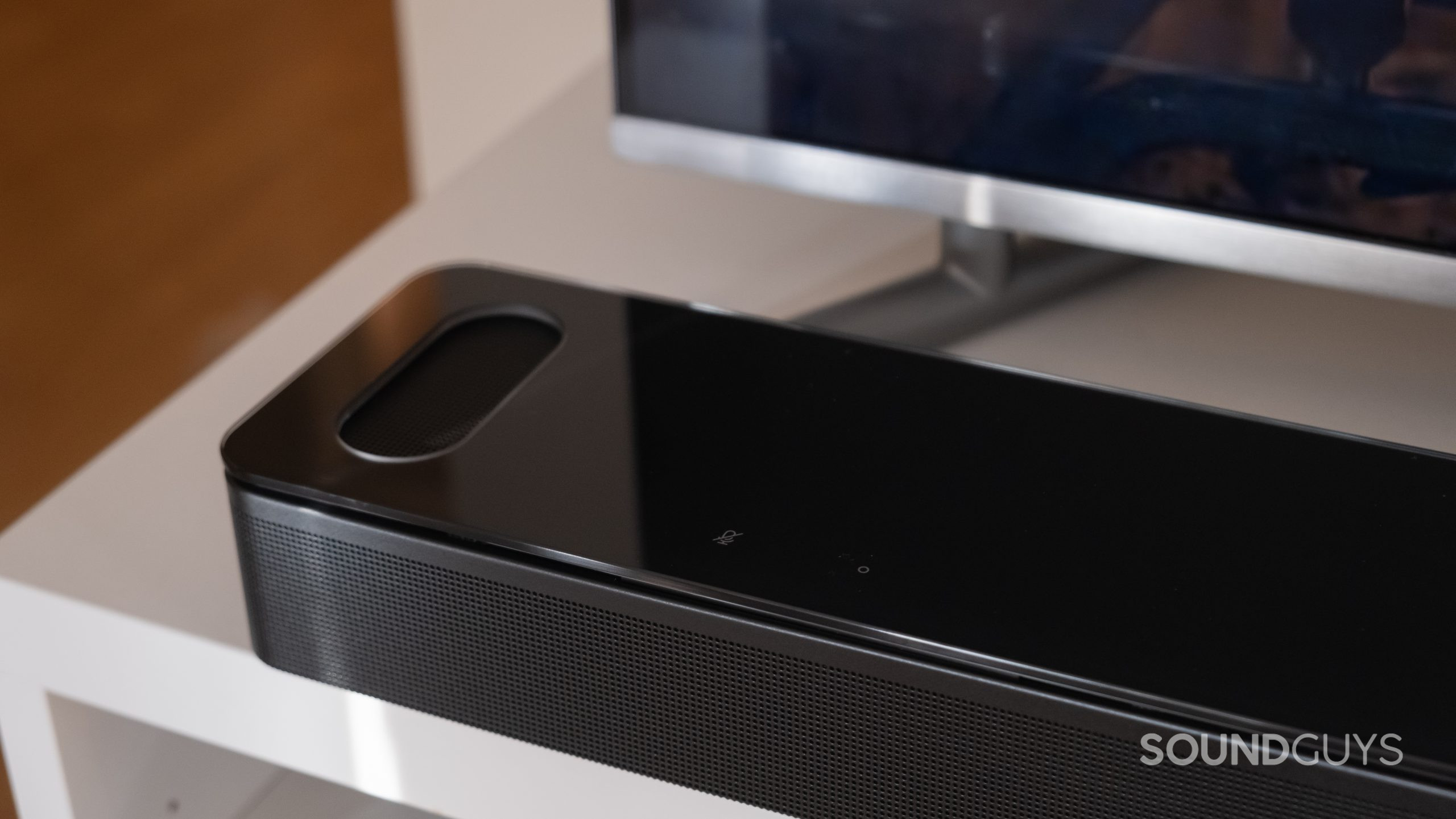 A close up of the Bose Smart Soundbar 900 on a TV stand with a TV in the background, showing the upfiring speaker.