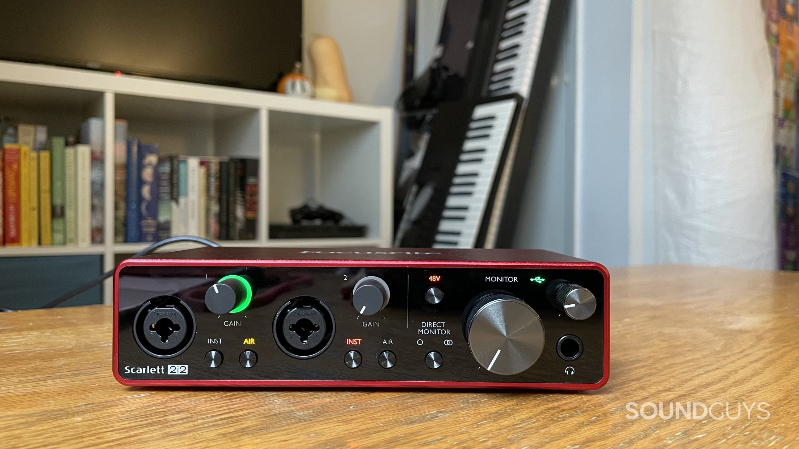 Focusrite Scarlett 2i2 on a table with a bookshelf and two electronic keyboards in the background. Several of the lights on the Scarlett 2i2 are on.