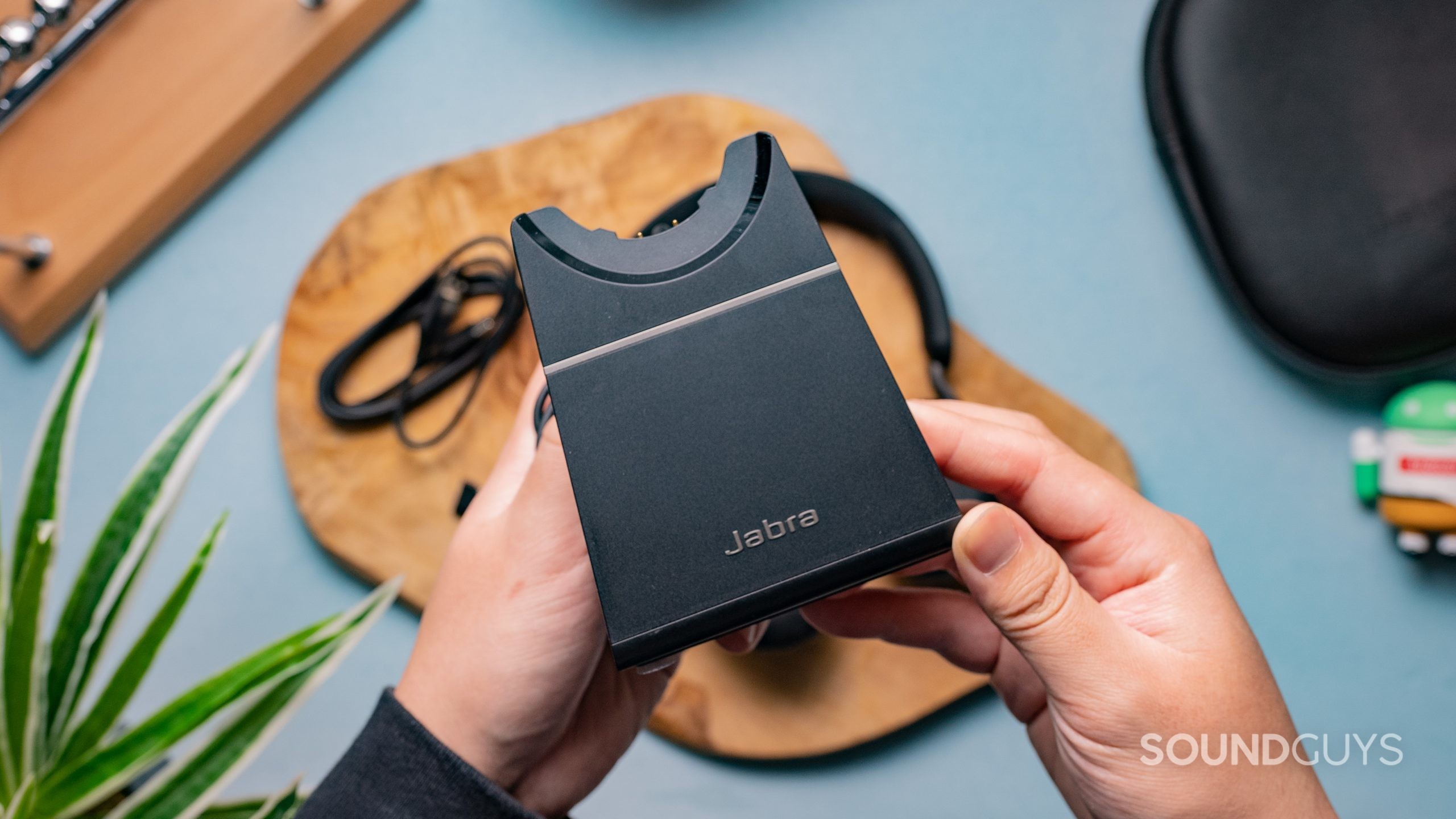 The Jabra Evolve2 65's charging cradle being held in someone's hands with the other accessories on the table behind it.