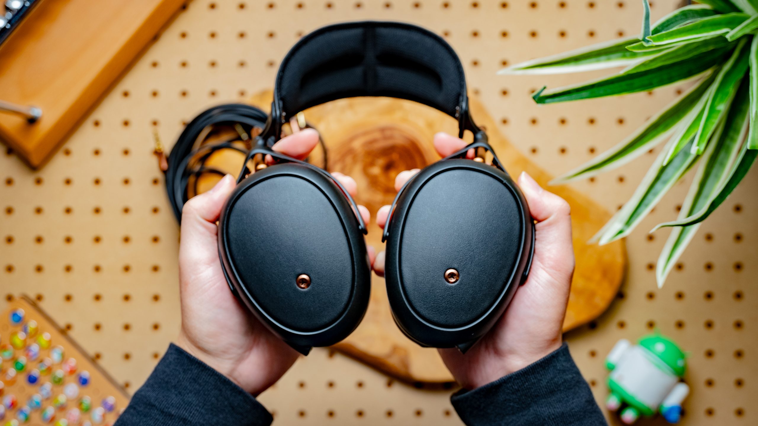 A photo of the Meze Audio Liric being held over pegboard, a plant, and wood.