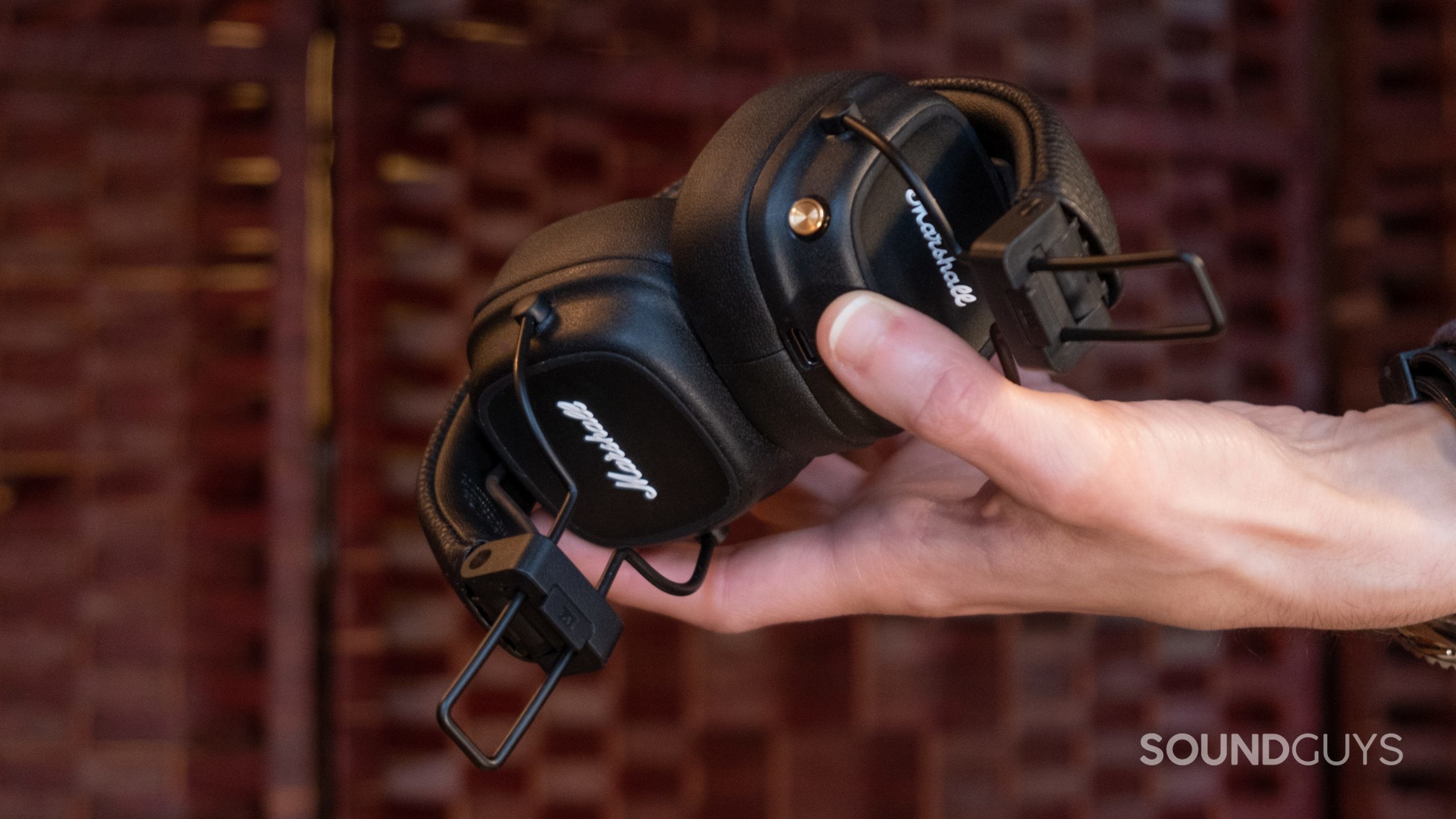 A hand holds up the folded down Marshall Major IV headphones in front of a rattan background.